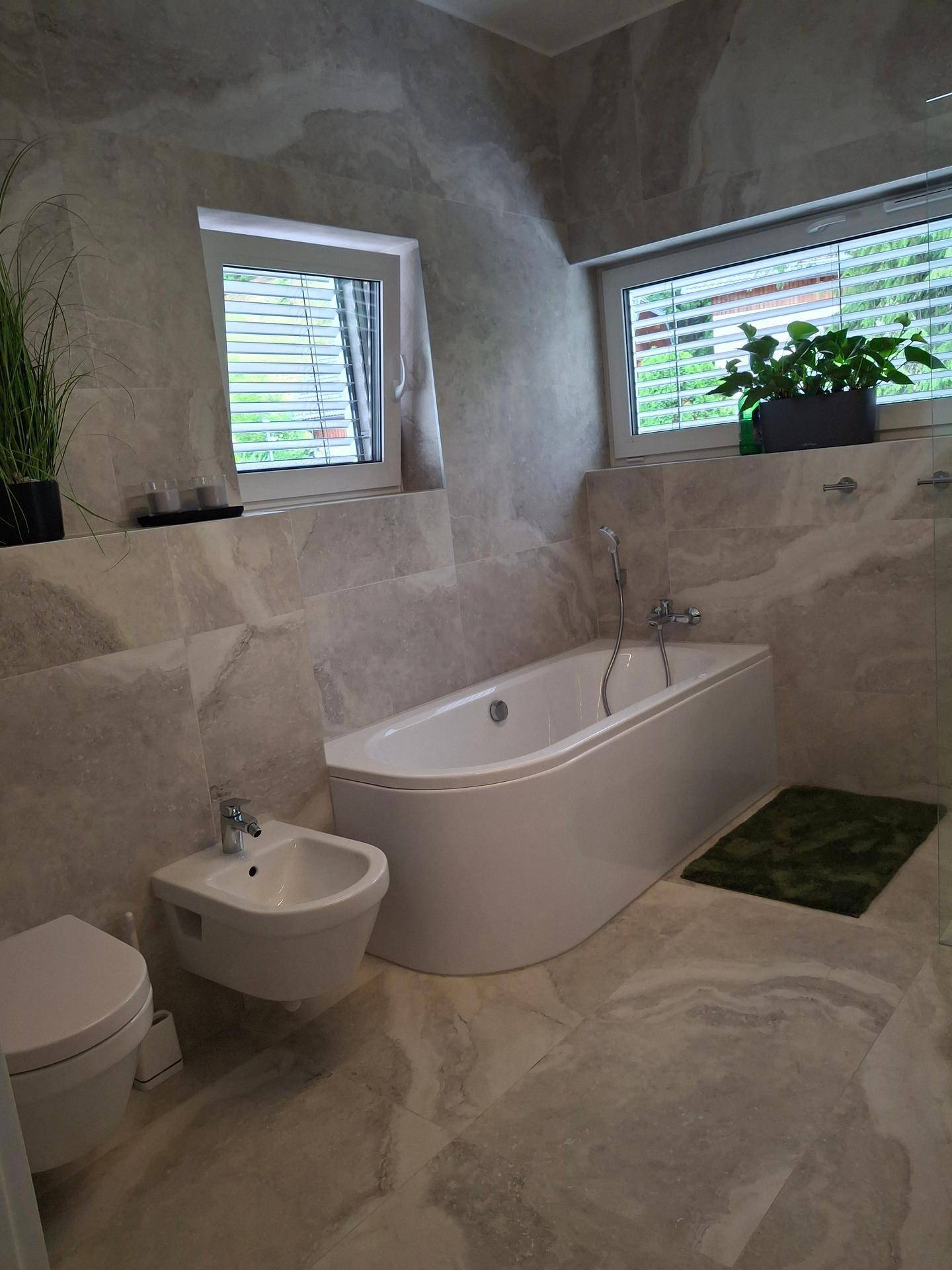 Modernes Badezimmer mit Badewanne, Bidet und hellen Fliesen, gut beleuchtet durch zwei Fenster.
