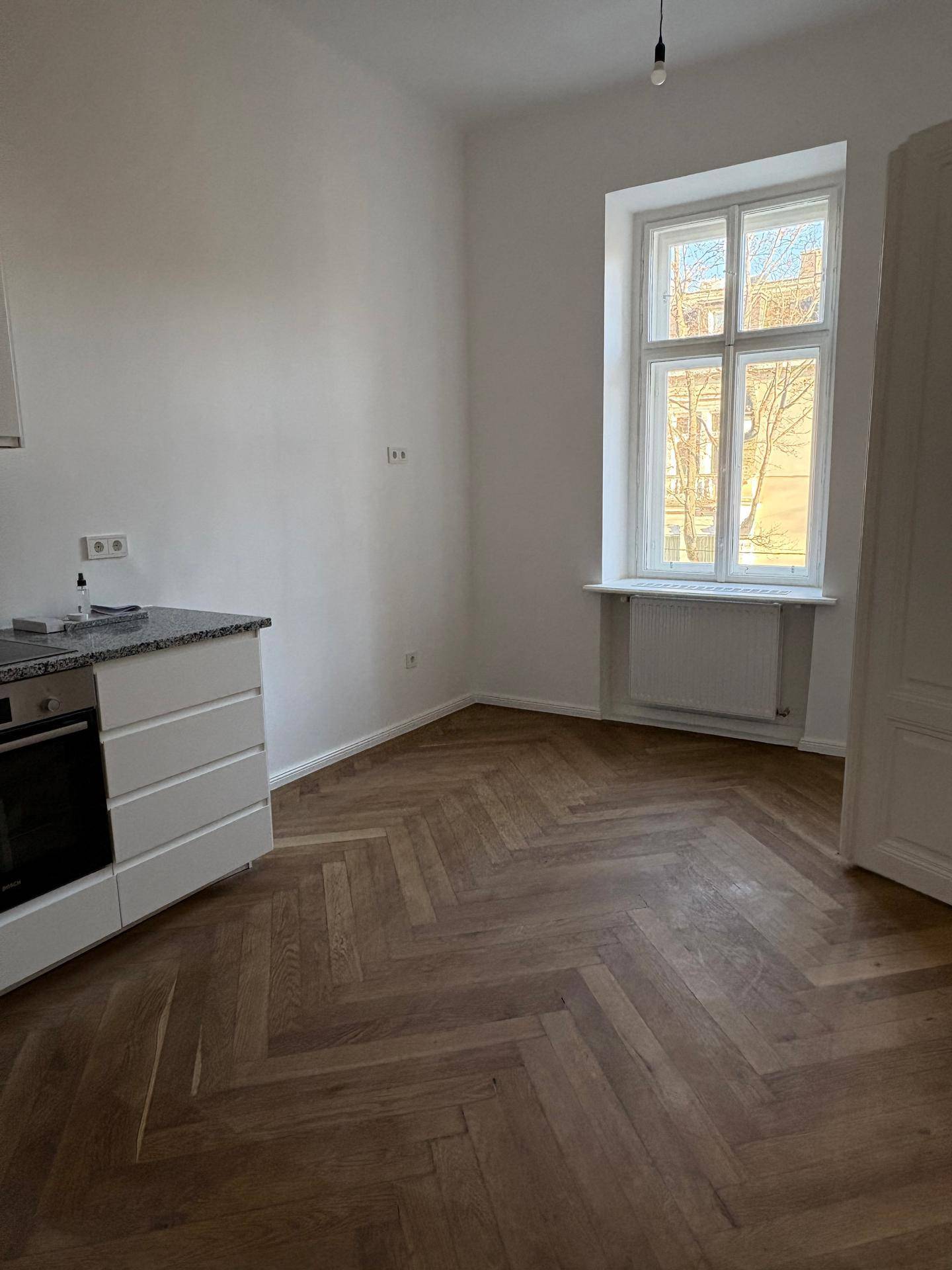 Heller Küchenbereich mit Fischgrätparkett, großem Fenster und Heizkörper unter der Fensterbank.