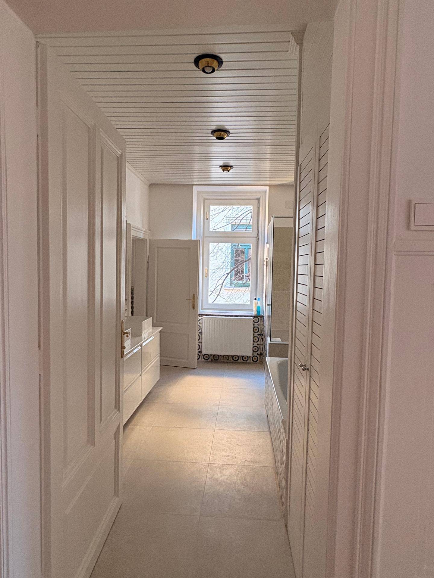 Langer Flur mit hellem Fliesenboden, der zu einem Badezimmer mit Fenster führt.