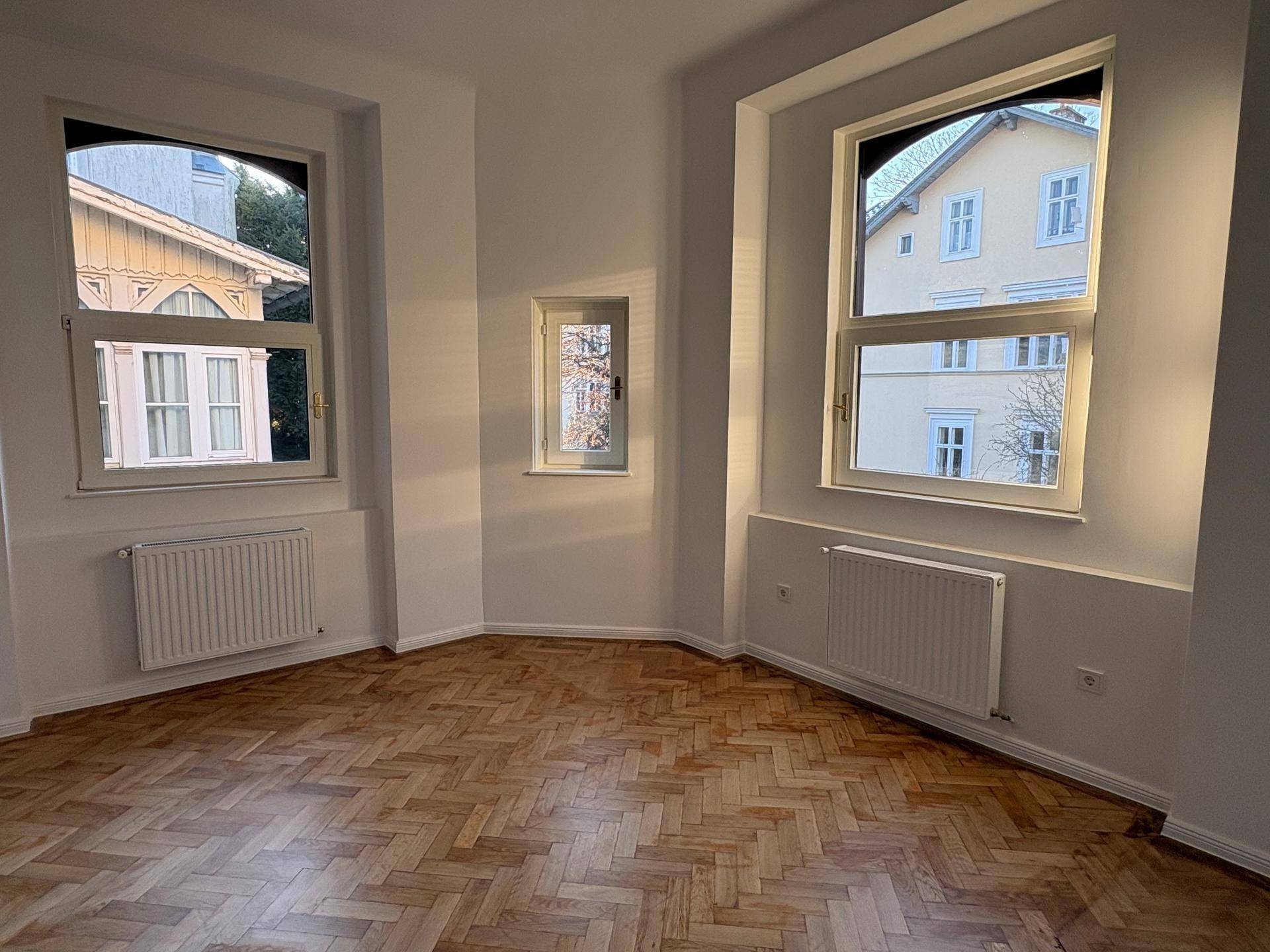 Helles Zimmer mit Fischgrätparkett und mehreren Fenstern, die viel Tageslicht hereinlassen.