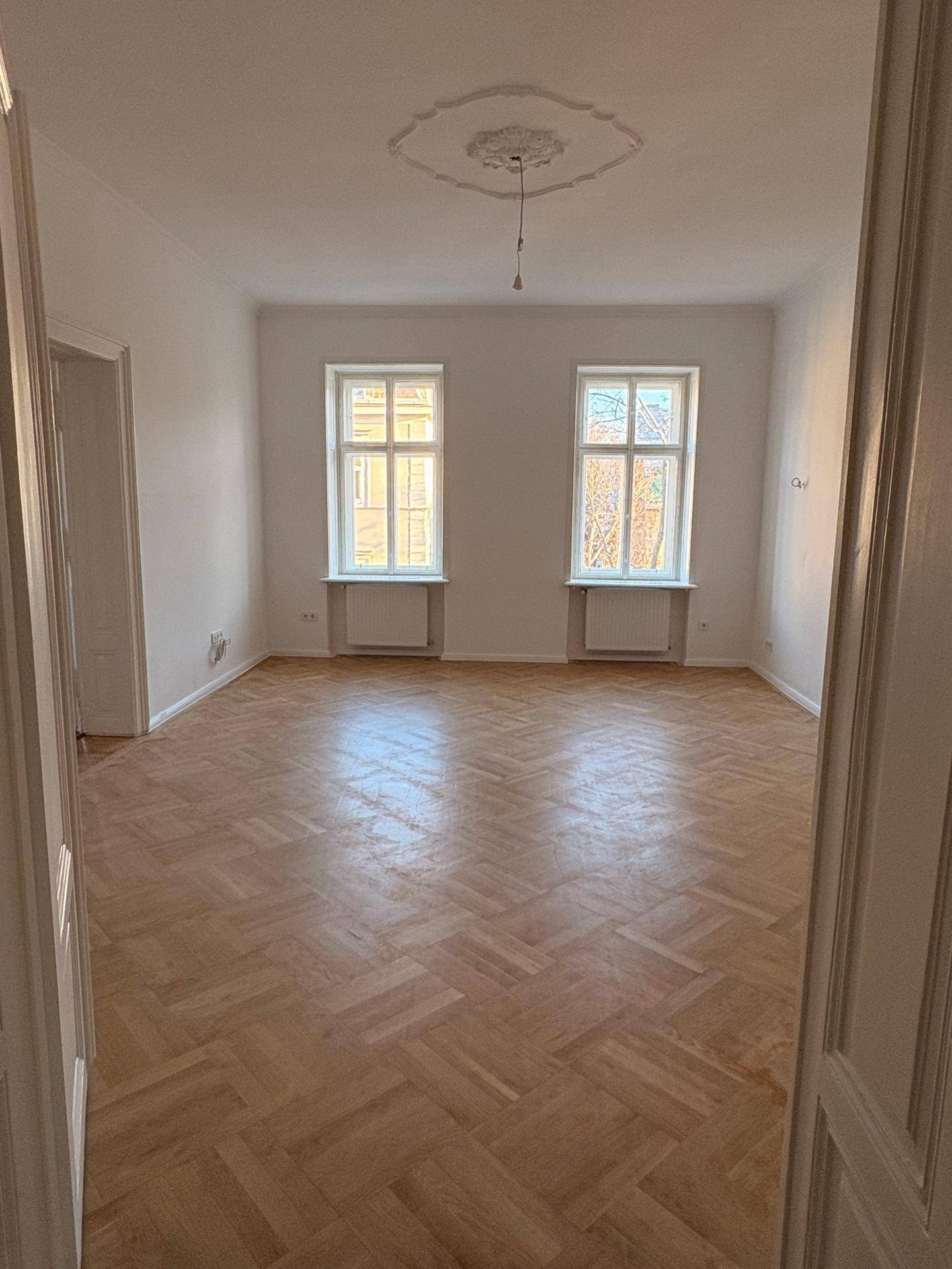 Geräumiges Zimmer mit Fischgrätparkett, hohen Decken und Stuckverzierung, durch zwei große Fenster erhellt.