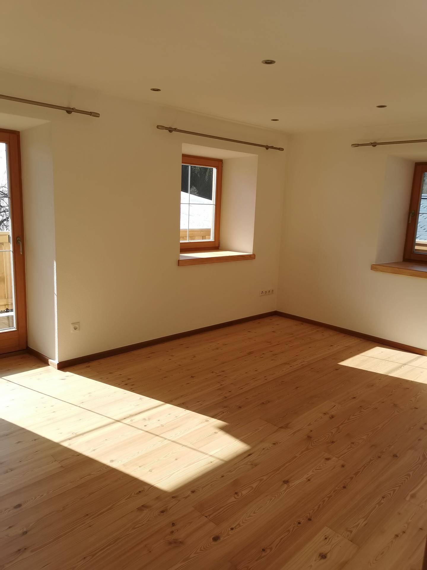Heller, leerer Raum mit Holzboden und mehreren Fenstern, die viel Tageslicht hereinlassen.