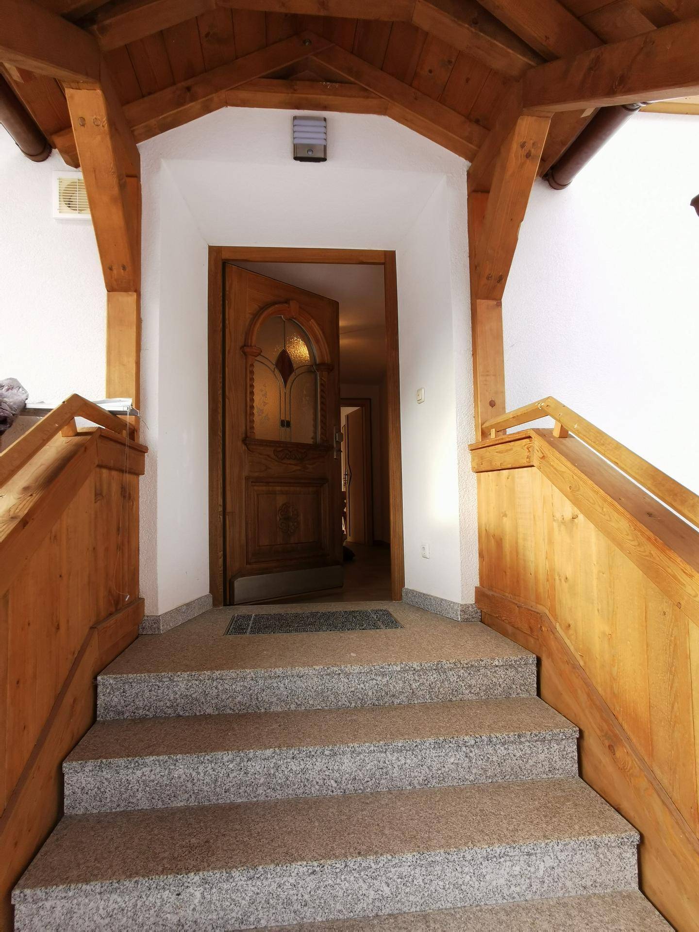 Überdachter Eingangsbereich mit Holzgeländer und Steintreppe, die zur Haustür führt.