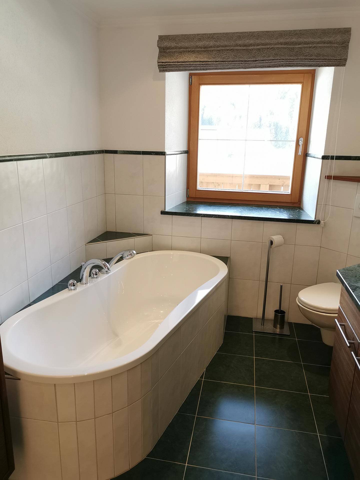 Badewanne und Toilette in einem Badezimmer mit Fenster und grünen Bodenfliesen.