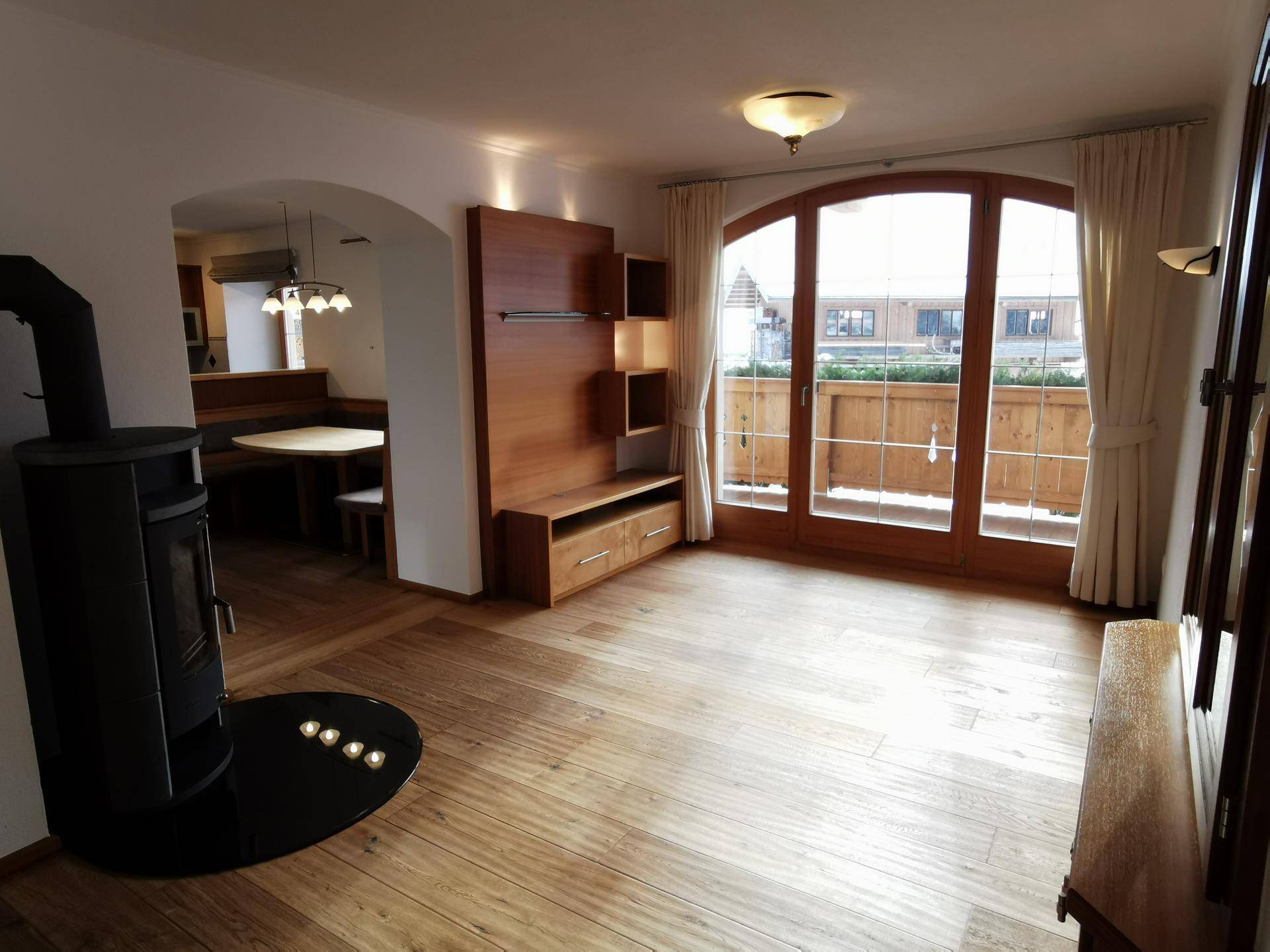 Wohnzimmer mit Holzofen, Holzboden und Zugang zu einem Balkon, im Hintergrund die Küche.