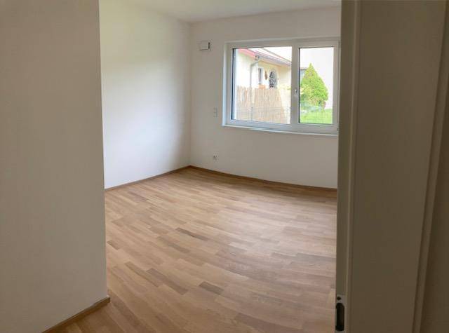 Leeres Zimmer mit Holzboden, weißen Wänden und einem großen Fenster mit Blick ins Grüne.