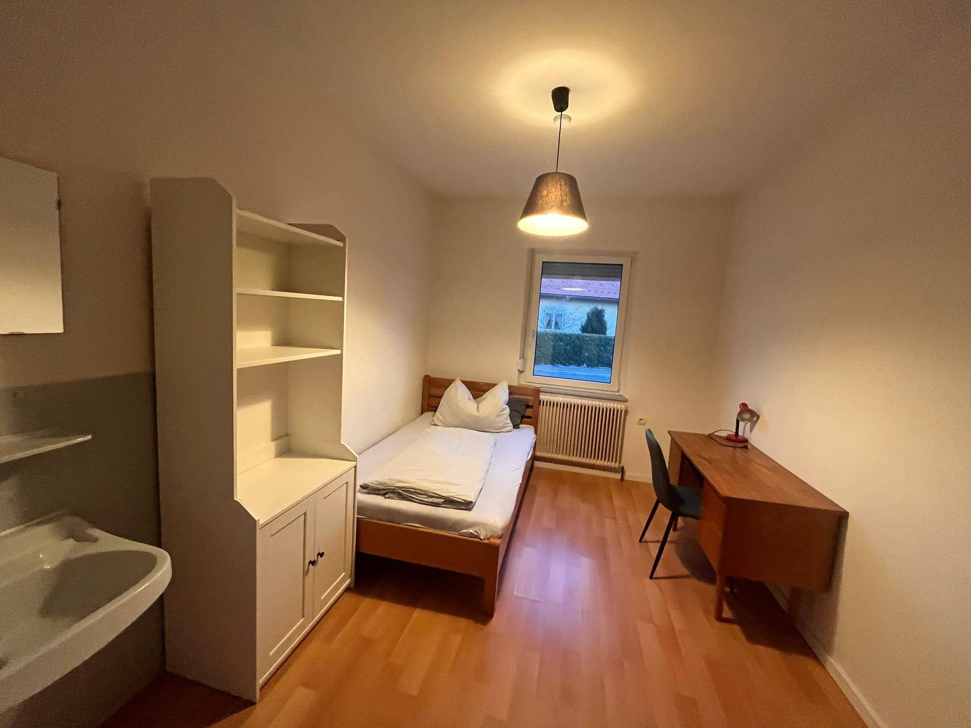Helles Schlafzimmer mit Einzelbett, Schreibtisch, Waschbecken und Fensterblick ins Grüne.