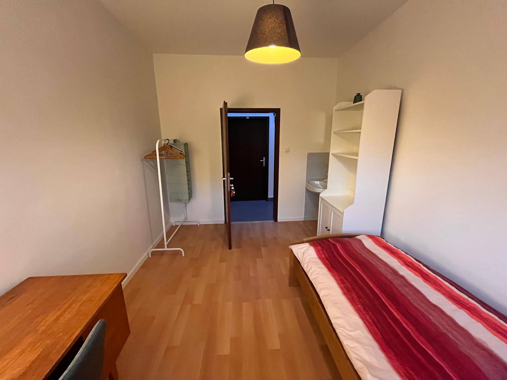 Schlafzimmer mit Einzelbett, Schreibtisch und integriertem Waschbecken in der Ecke.