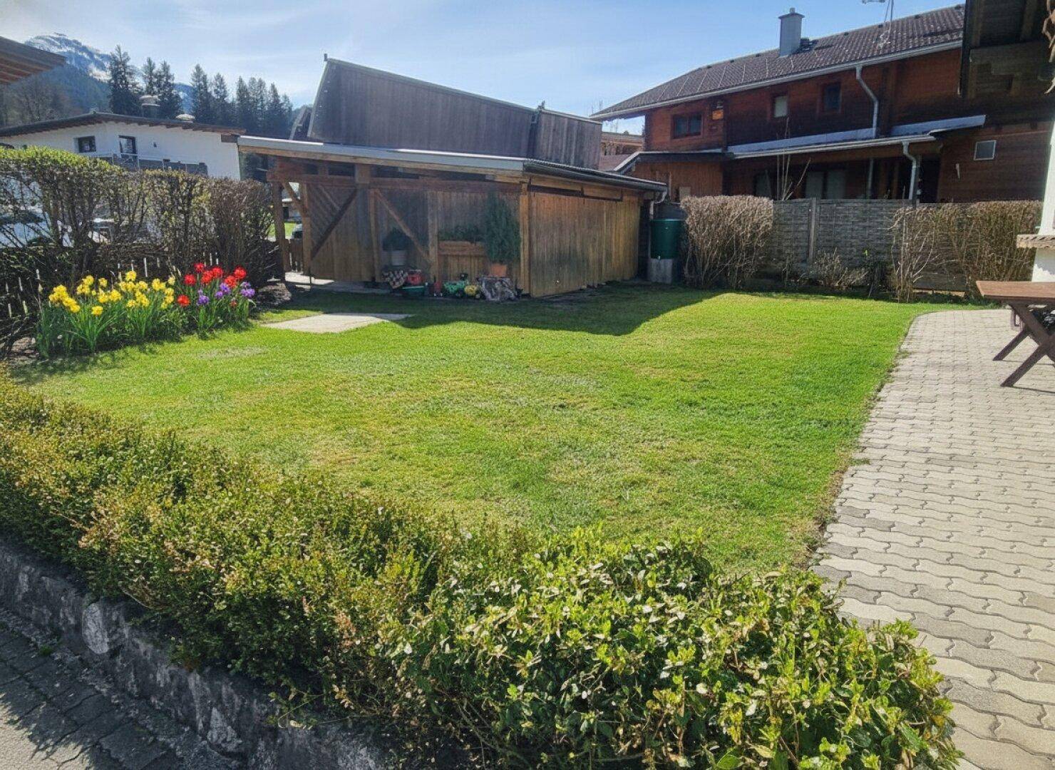 Gepflegter Gartenbereich mit Rasenfläche, blühenden Blumenbeeten und einem Holzschuppen.
