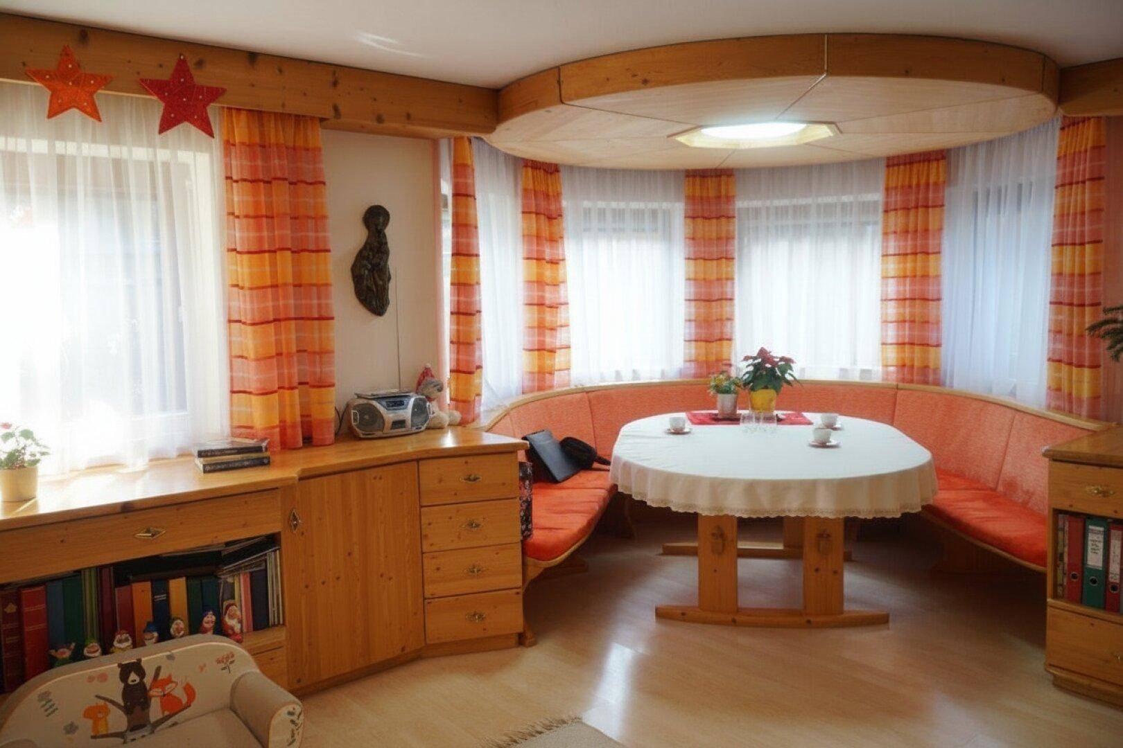 Heller Essbereich mit runder Holzbank, Esstisch und Fenstern mit farbigen Vorhängen.