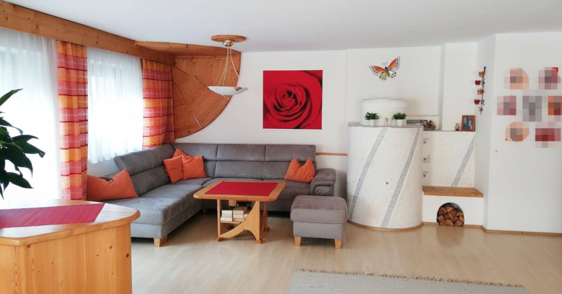 Gemütliches Wohnzimmer mit Ecksofa, traditionellem Kachelofen und Holzelementen an der Decke.