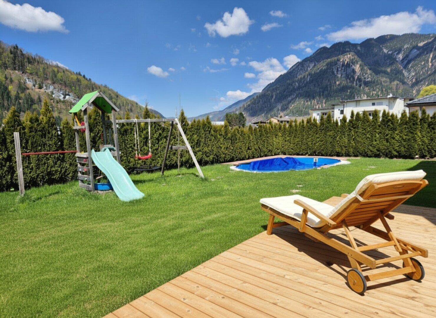 Großer Garten mit Spielplatz, Pool und Holzterrasse, umgeben von einer Hecke und Bergen.