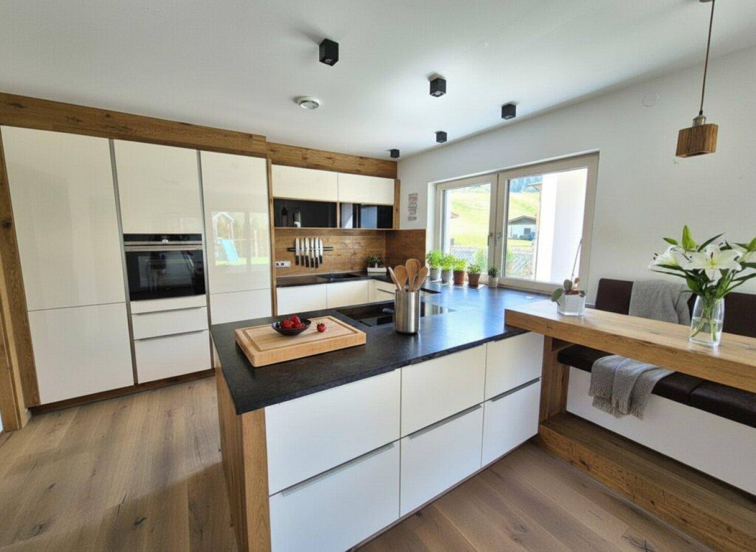 Moderne Küche mit Kochinsel, weißen Hochglanzfronten, Holzelementen und Essbereich am Fenster.