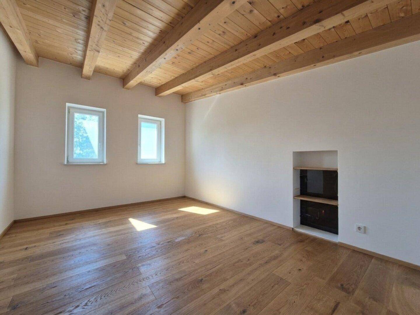 Lichtdurchflutetes Zimmer mit Holzbalkendecke, Holzboden und zwei Fenstern.