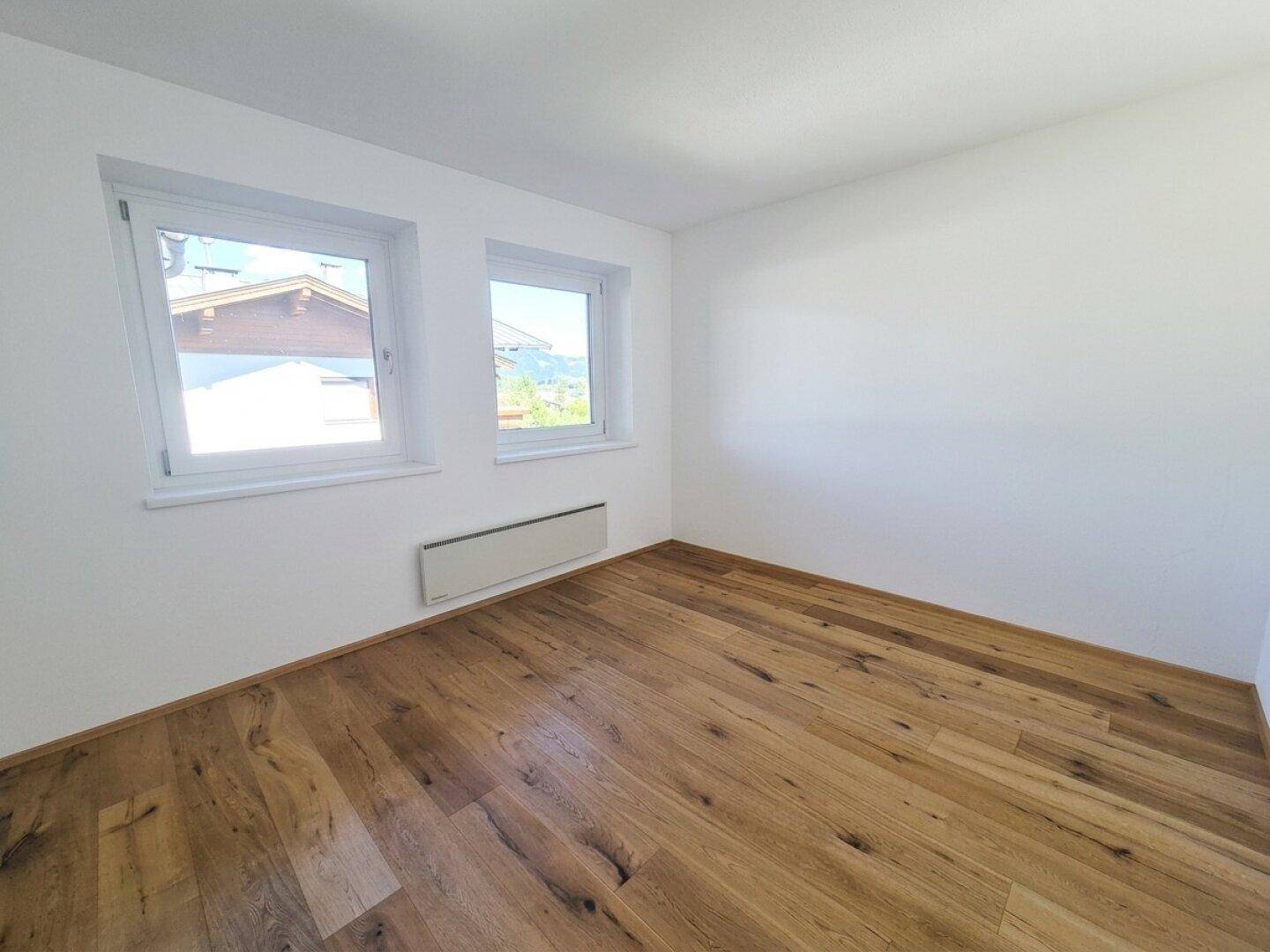 Helles leeres Zimmer mit Holzboden und zwei Fenstern.