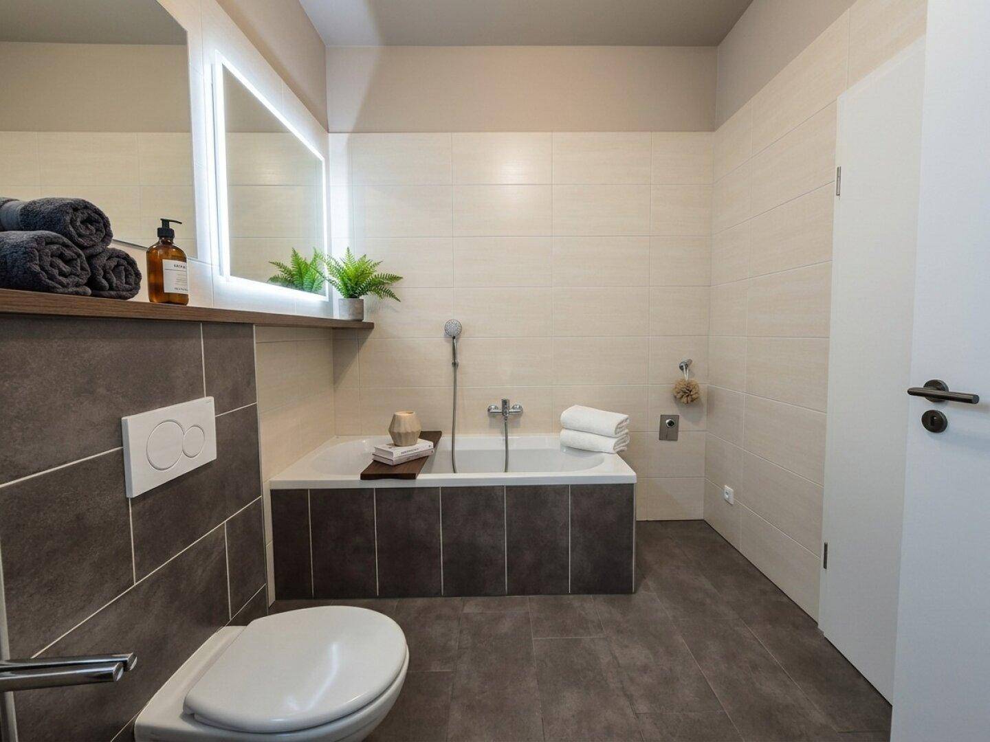 Modernes Badezimmer mit Badewanne, Wand-WC und großem beleuchteten Spiegel.