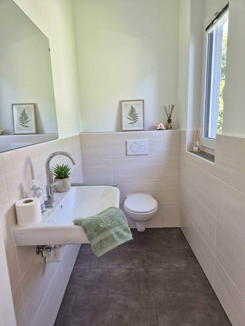 Modernes Gäste-WC mit Wand-WC, kleinem Waschbecken und Fenster für natürliche Belüftung.