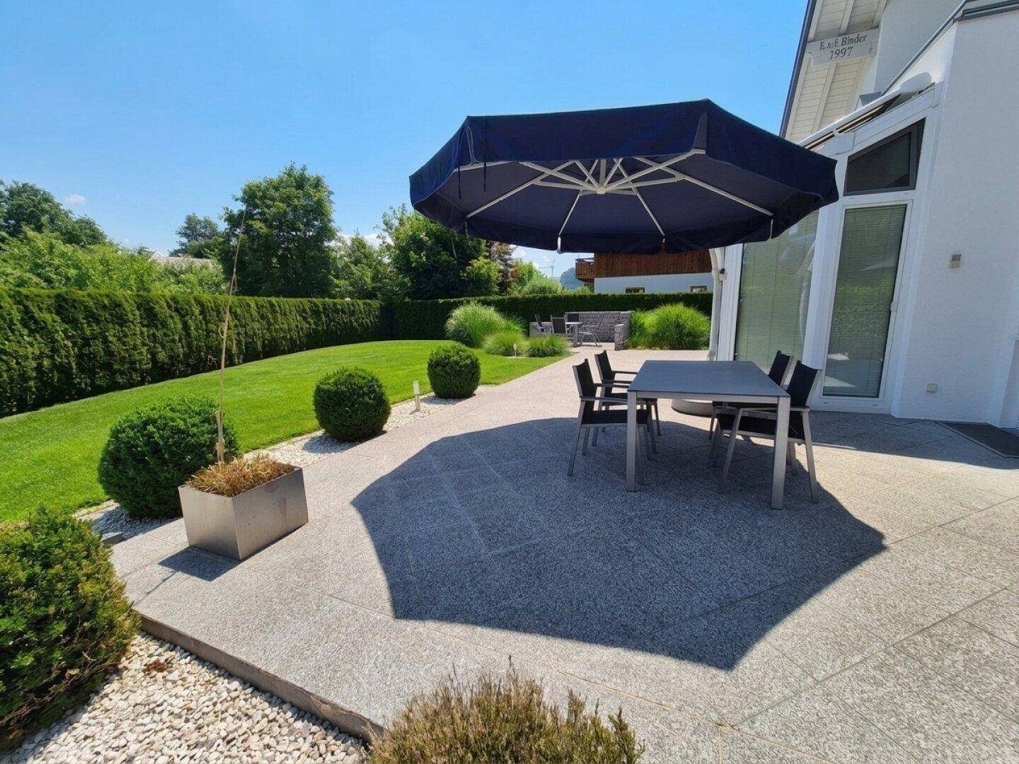 Gemütliche Terrasse mit Esstisch und Sonnenschirm, umgeben von einem gepflegten Garten und Hecken.