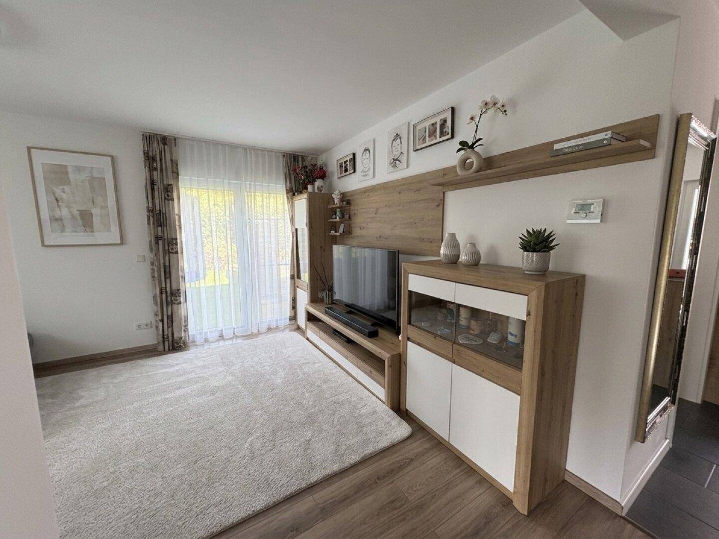 Helles Wohnzimmer mit modernem TV-Möbel, Teppichboden und Zugang zum Außenbereich.