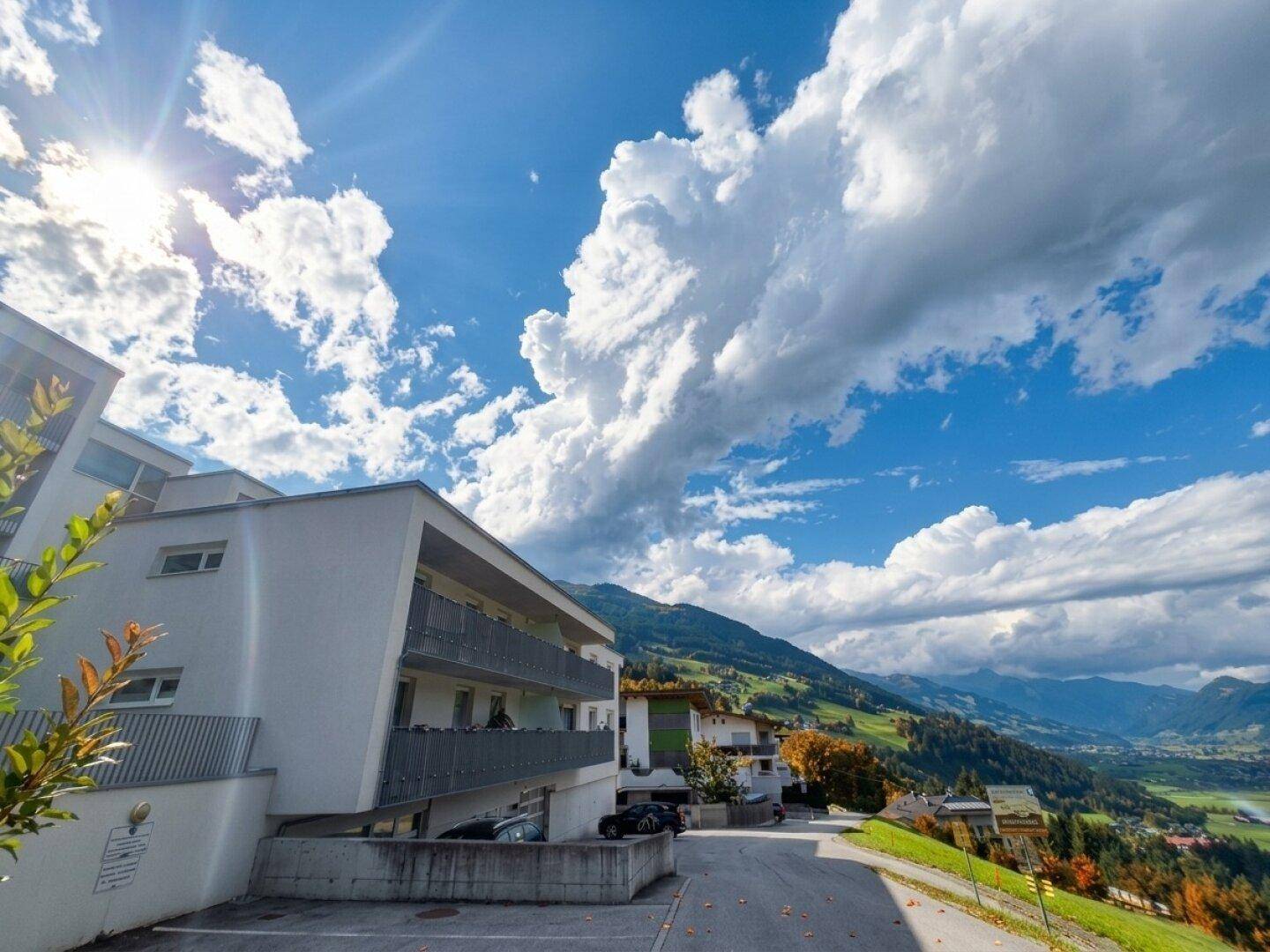 Modernes Mehrfamilienhaus in Hanglage mit Balkonen und Bergblick unter blauem Himmel.