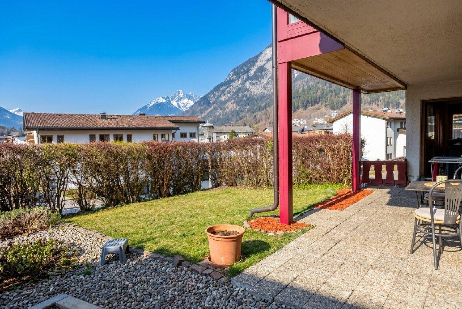 Sonniger Gartenbereich mit Terrasse, Rasenfläche und Hecke, umgeben von Bergen.