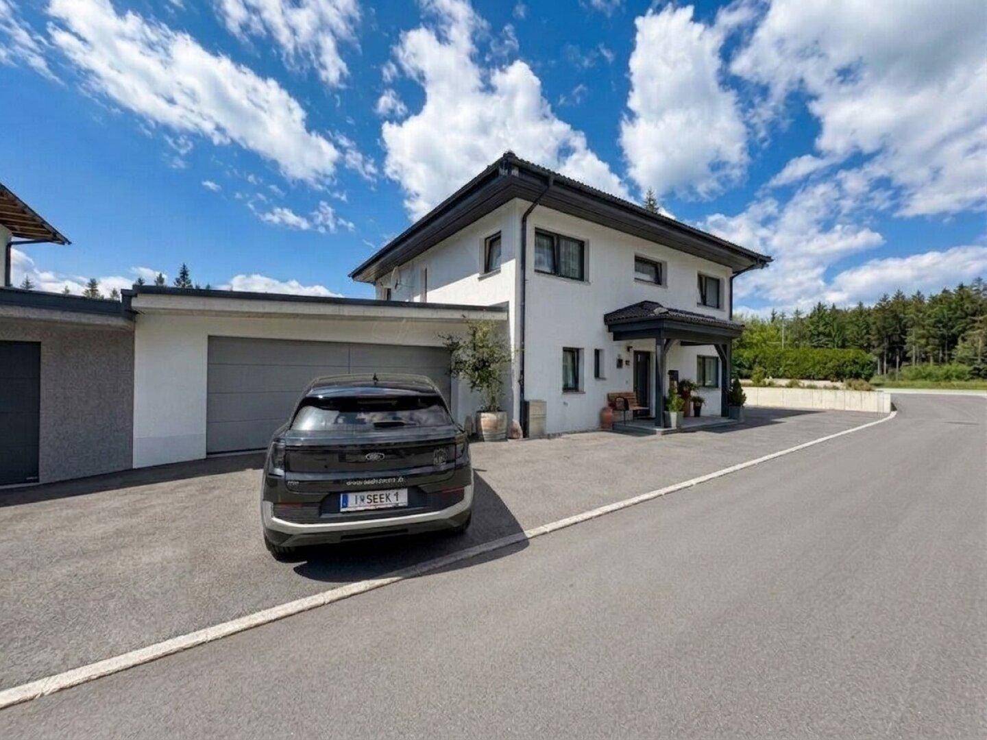 Moderne Hausfassade mit Doppelgarage und breiter Einfahrt, umgeben von einer gepflegten Landschaft und Wald.