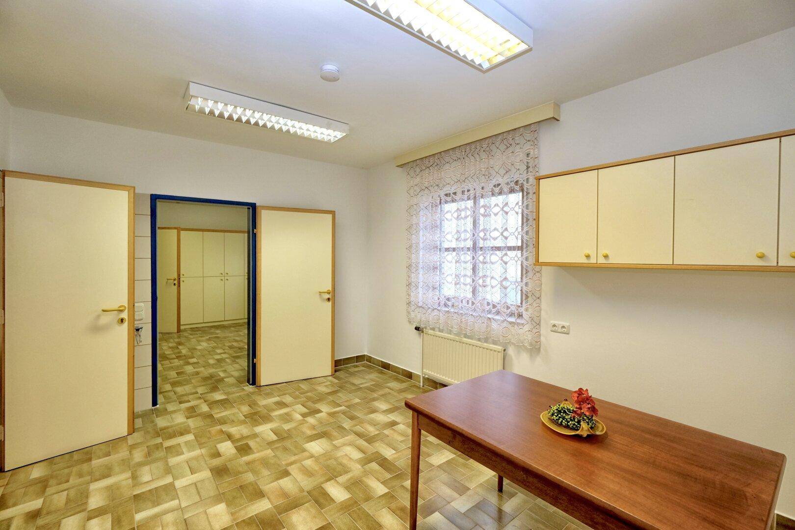 Kleinerer Büroraum mit einem Holztisch, Hängeschränken und einem Fenster mit Vorhang.