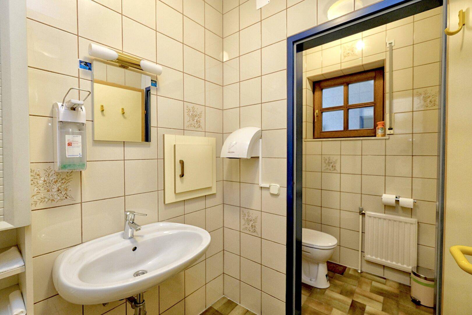 Gefliestes Badezimmer mit Waschbecken, Toilette, Fenster und Haltegriffen für Barrierefreiheit.