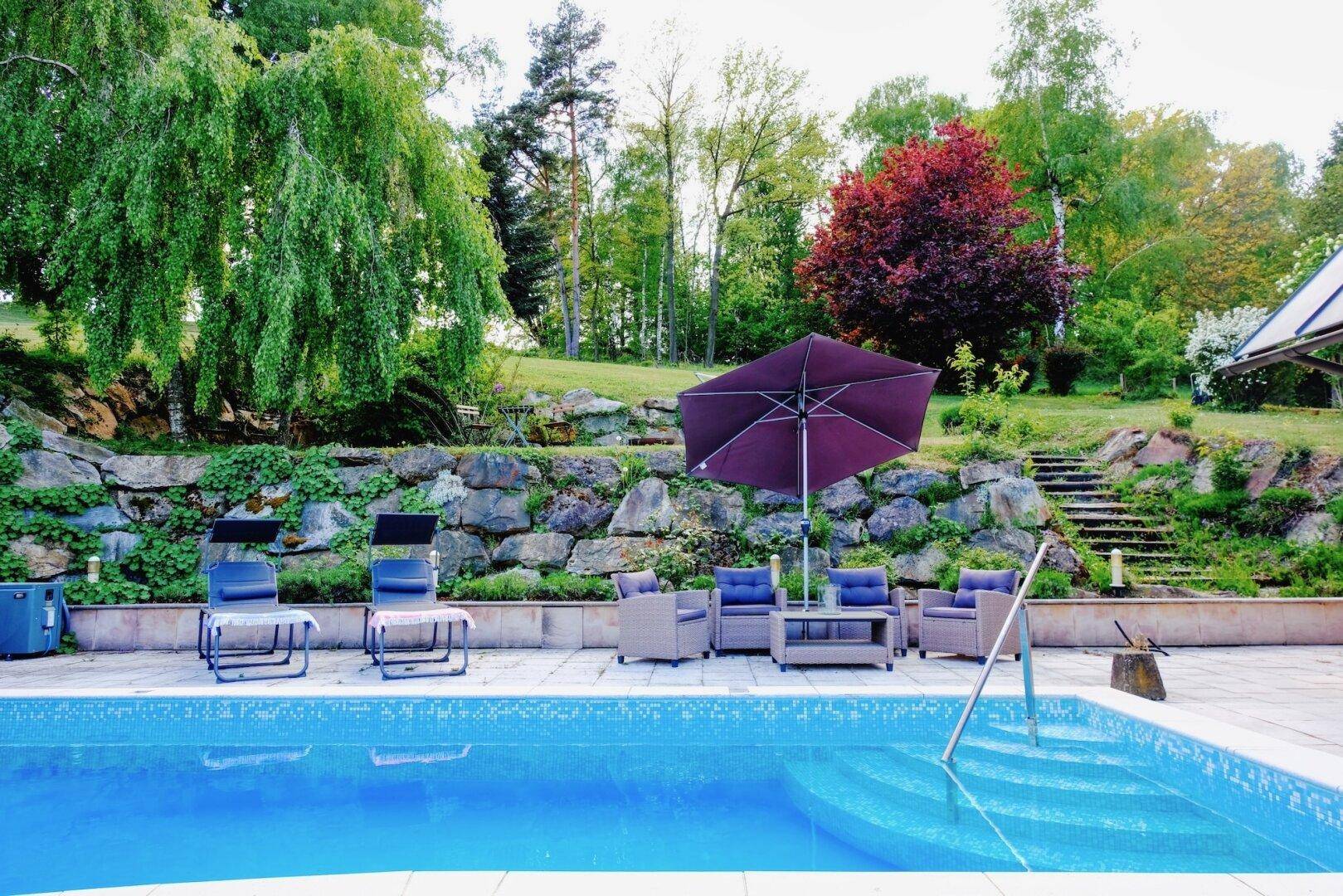 Poolbereich mit bequemen Gartenmöbeln, Sonnenschirm und einer Steinmauer im Hintergrund.
