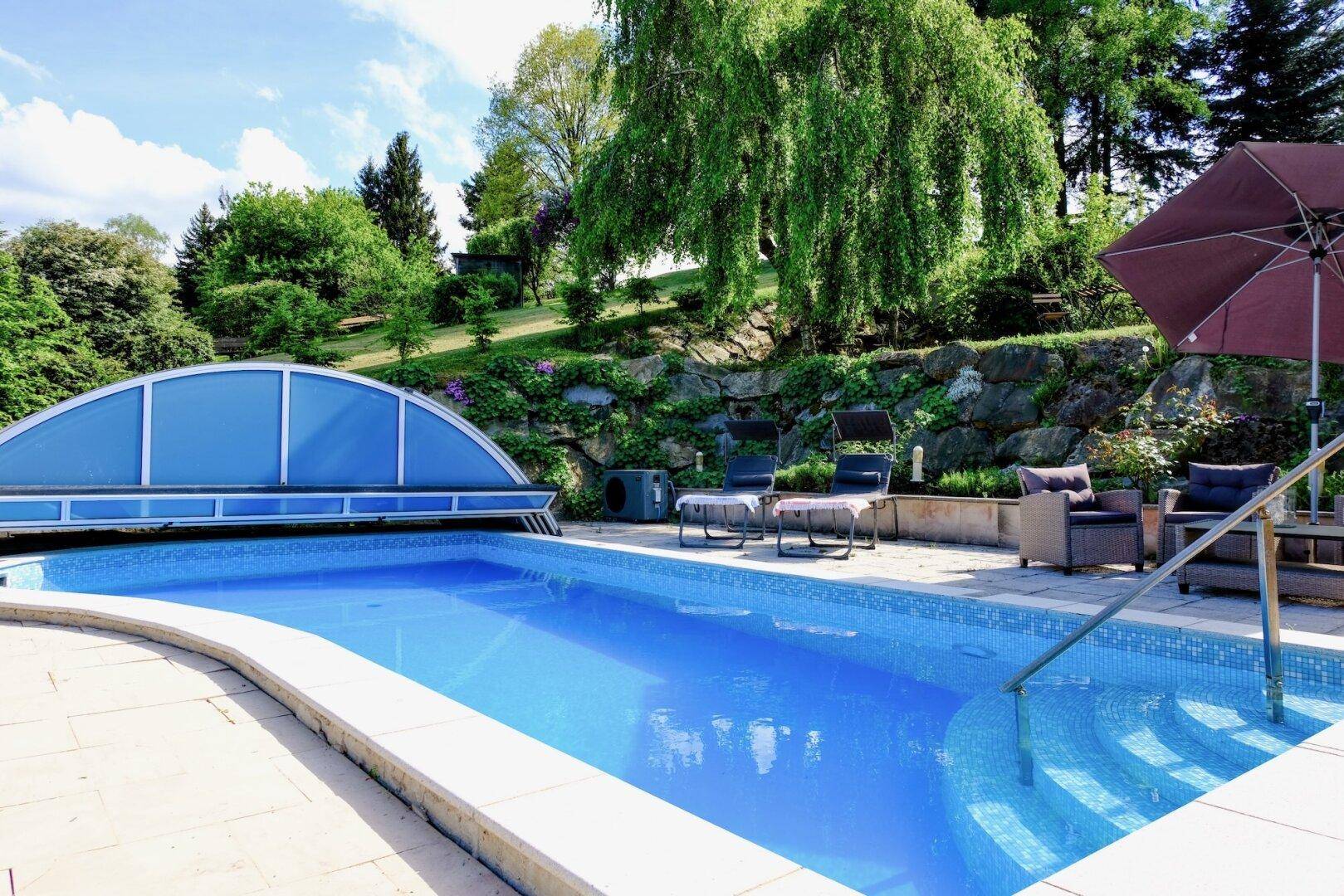 Blauer Swimmingpool mit römischer Treppe, umgeben von einer gepflasterten Terrasse und Grünflächen.
