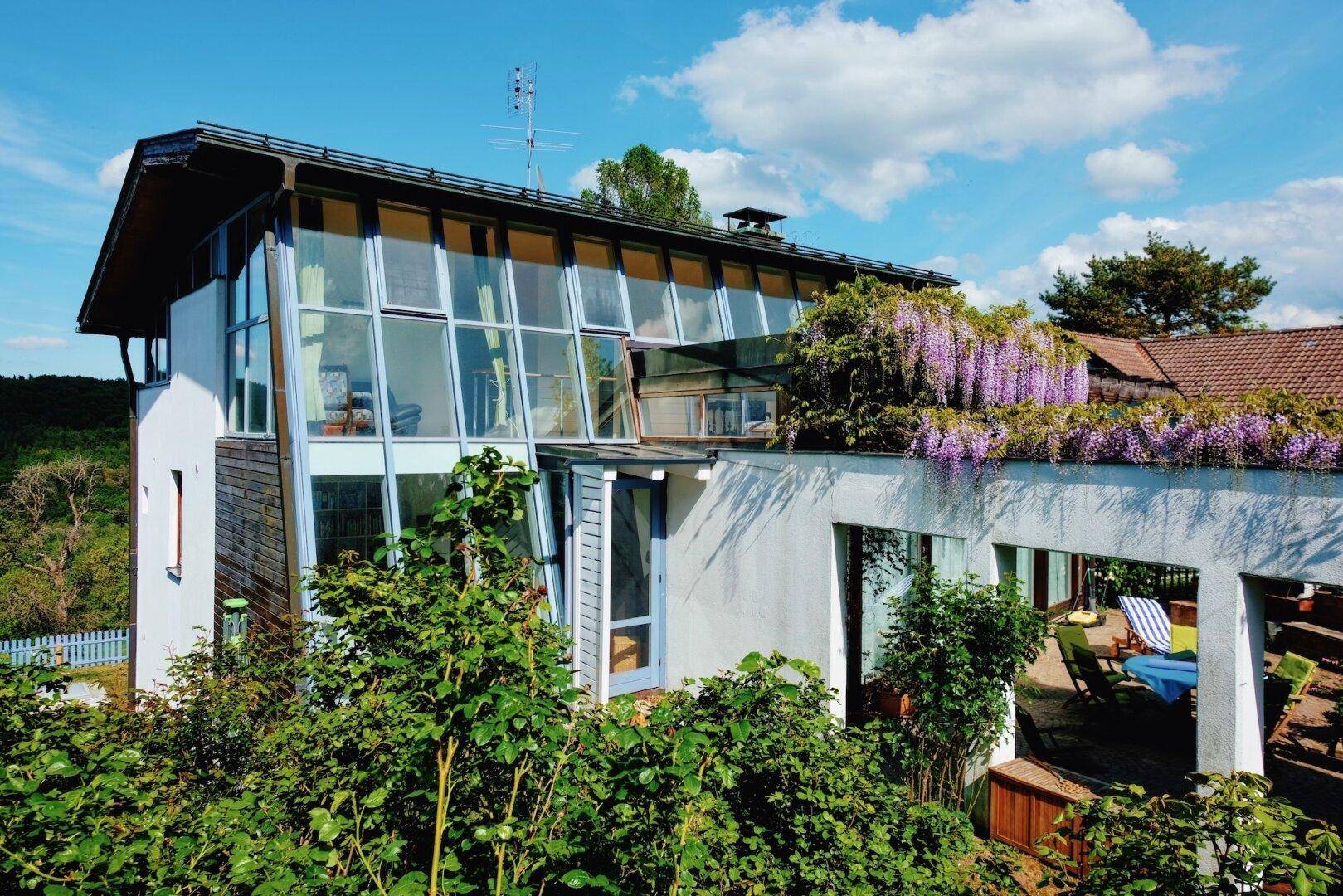 Modernes Haus mit großer Glasfassade, blühenden Pflanzen und grüner Umgebung.