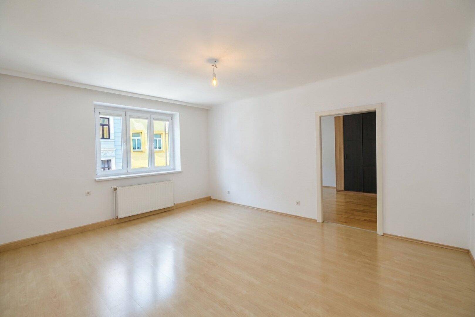 Leeres Wohnzimmer mit Holzboden, weißen Wänden und großen Fenstern.