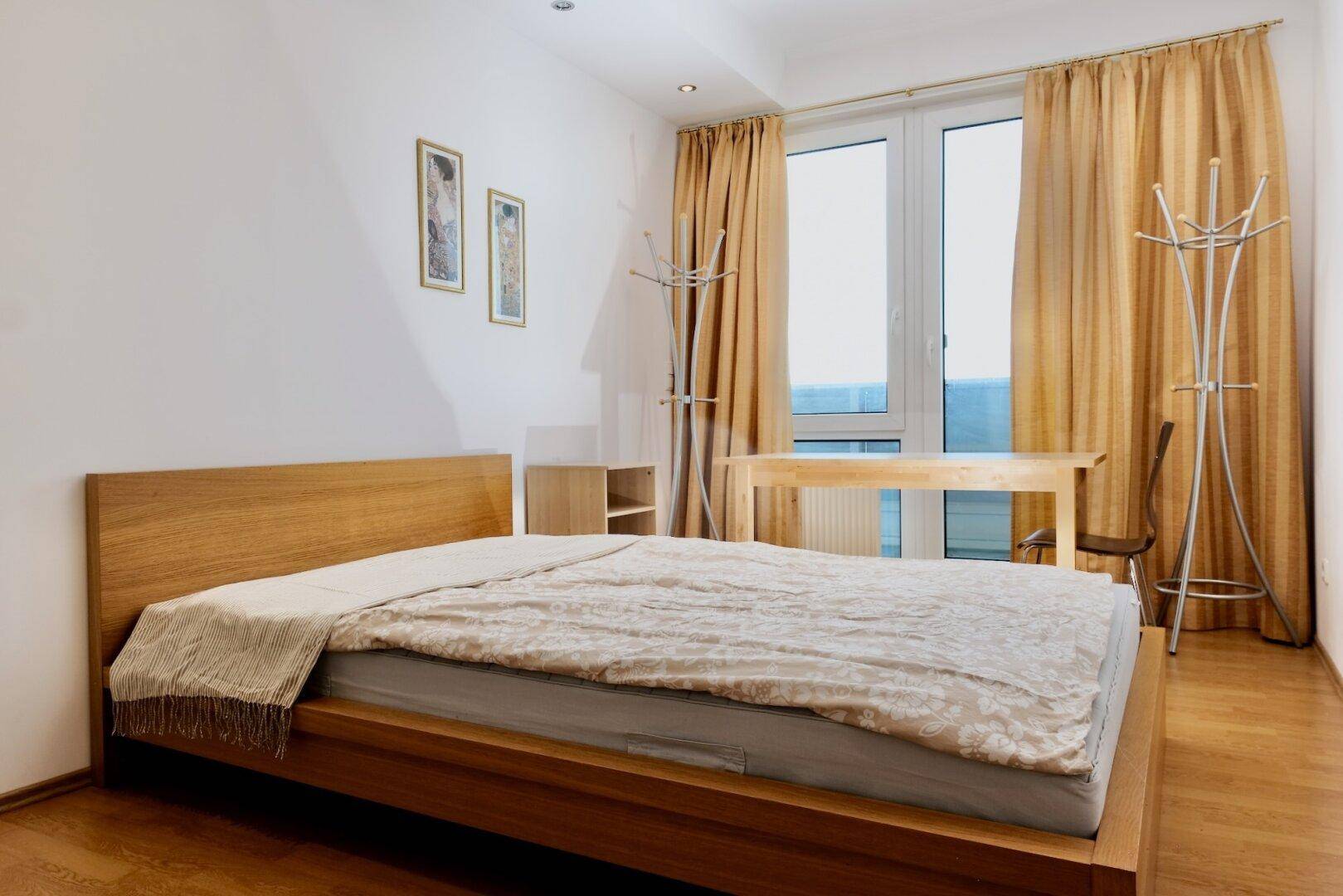 Schlafzimmer mit Holzbett, Parkettboden und großen Fenstern mit Vorhängen.
