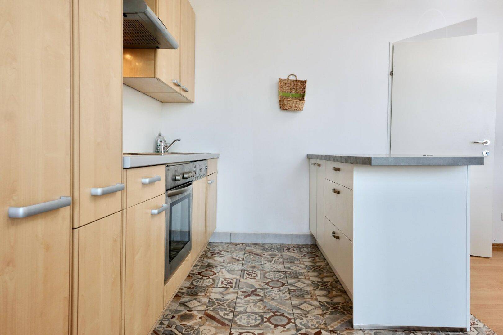 Funktionale Küche mit hellen Holzschränken, integriertem Backofen und gemusterten Fliesenboden.
