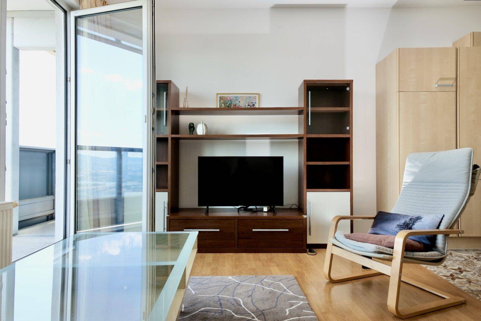 Gemütliches Wohnzimmer mit TV-Möbeln, Holzboden und Zugang zum Balkon.