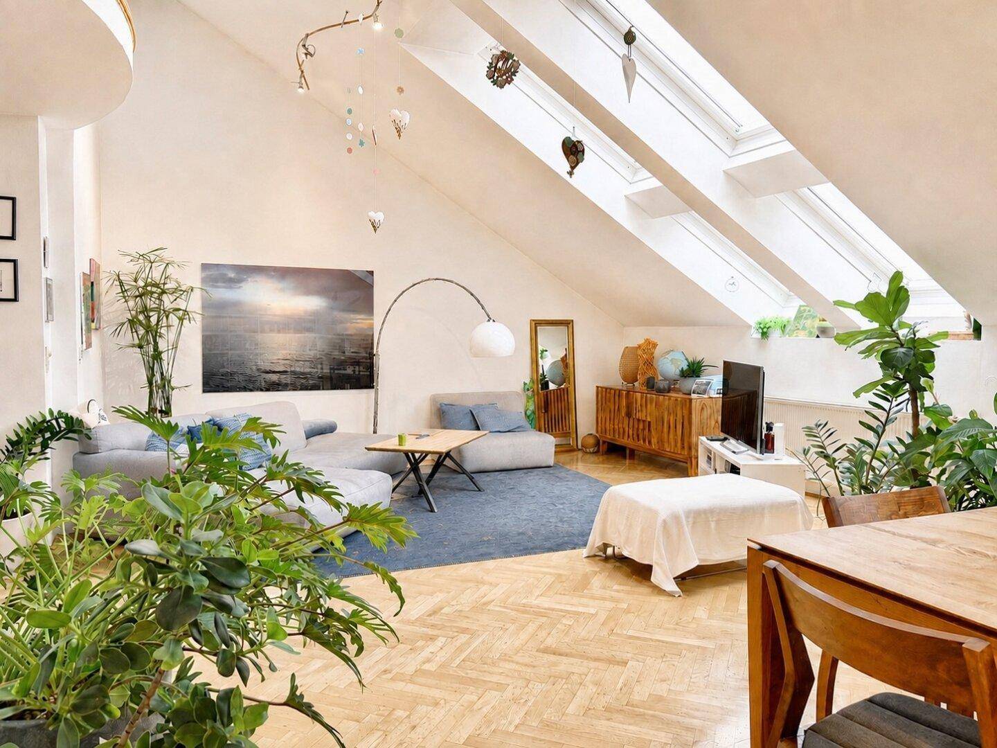 Gemütliches Wohnzimmer mit bequemer Couch, Teppich und vielen Pflanzen unter den Dachfenstern.
