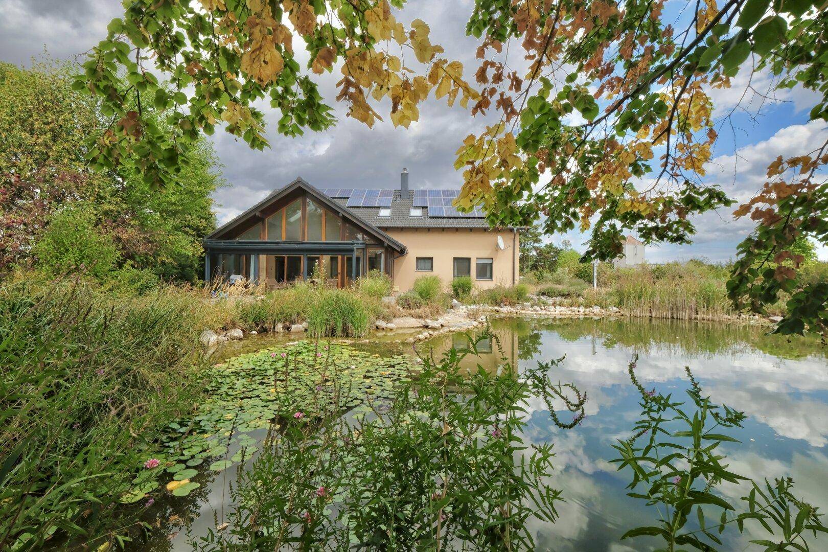 Haus mit Solaranlage am Teich, umgeben von üppiger Vegetation und herbstlichem Laub.