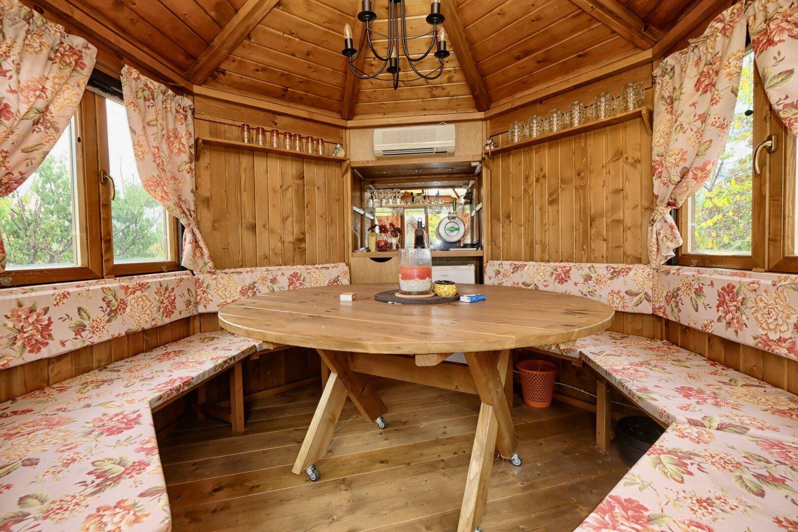 Gemütliches Gartenhaus aus Holz mit Sitzbänken und einem runden Tisch, umgeben von Natur.