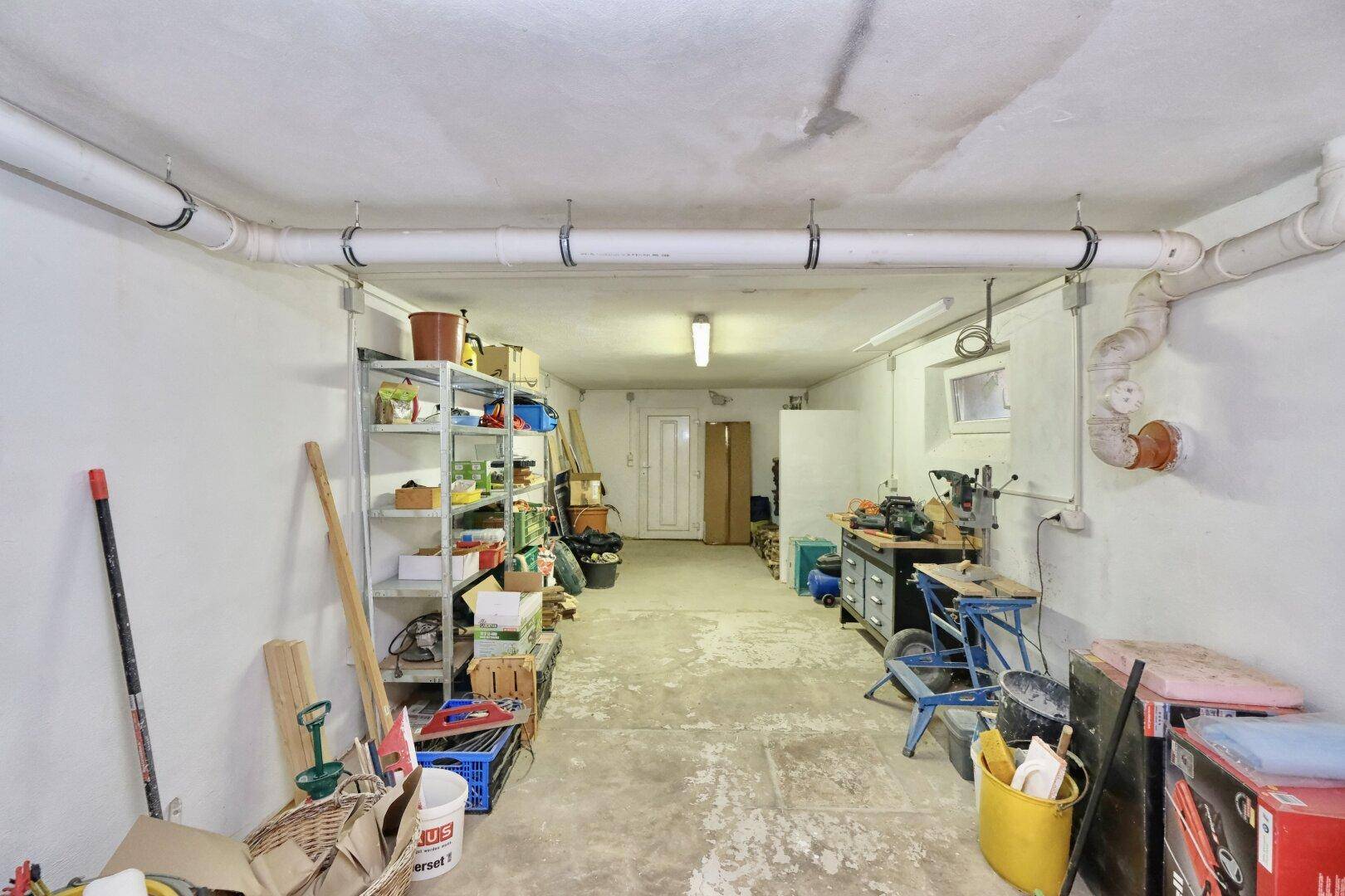 Geräumiger Kellerraum mit Regalen, Werkbank und sichtbaren Rohren, ideal als Lager oder Werkstatt.
