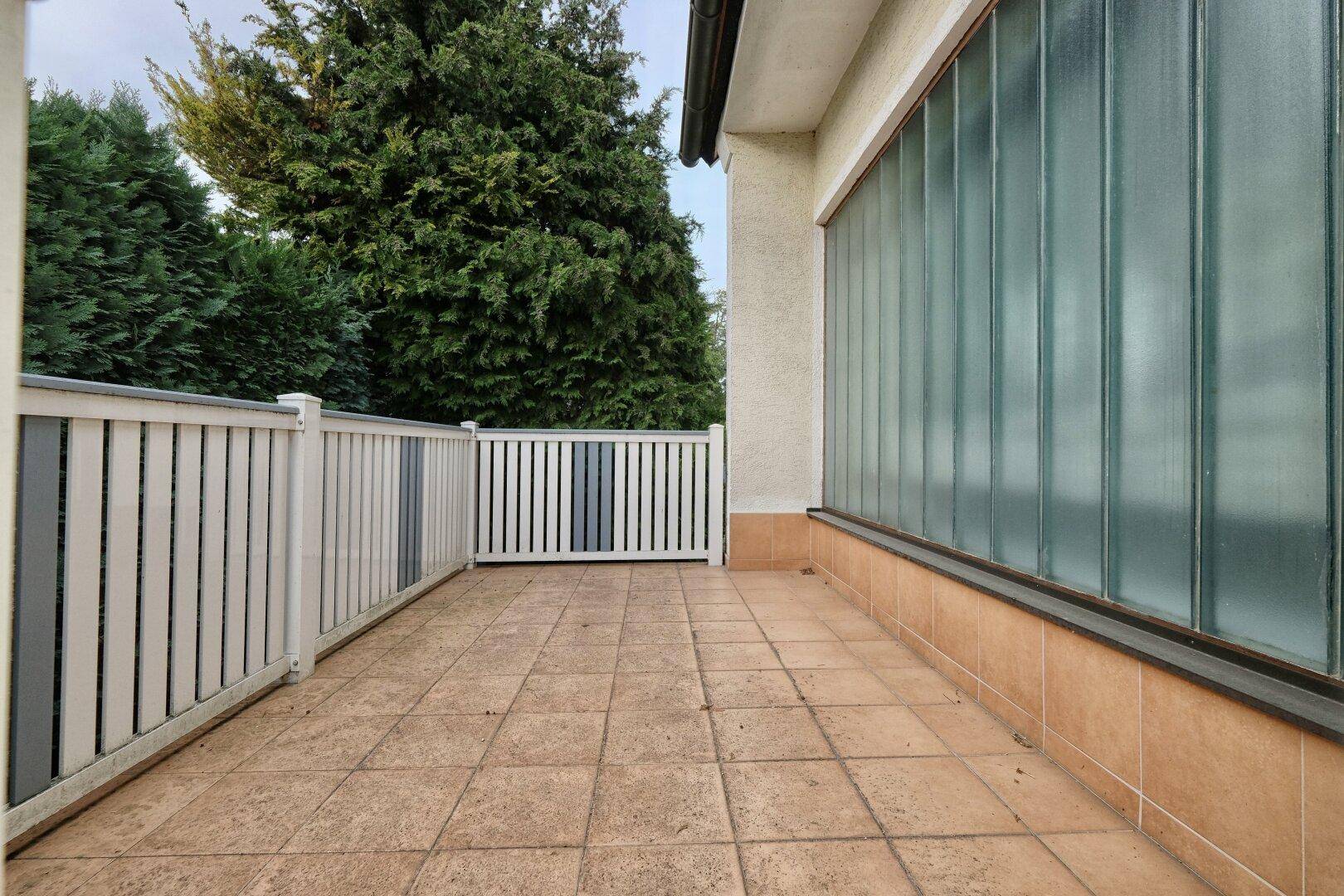 Geflieste Terrasse mit weißem Geländer und Blick auf die umliegende grüne Bepflanzung.