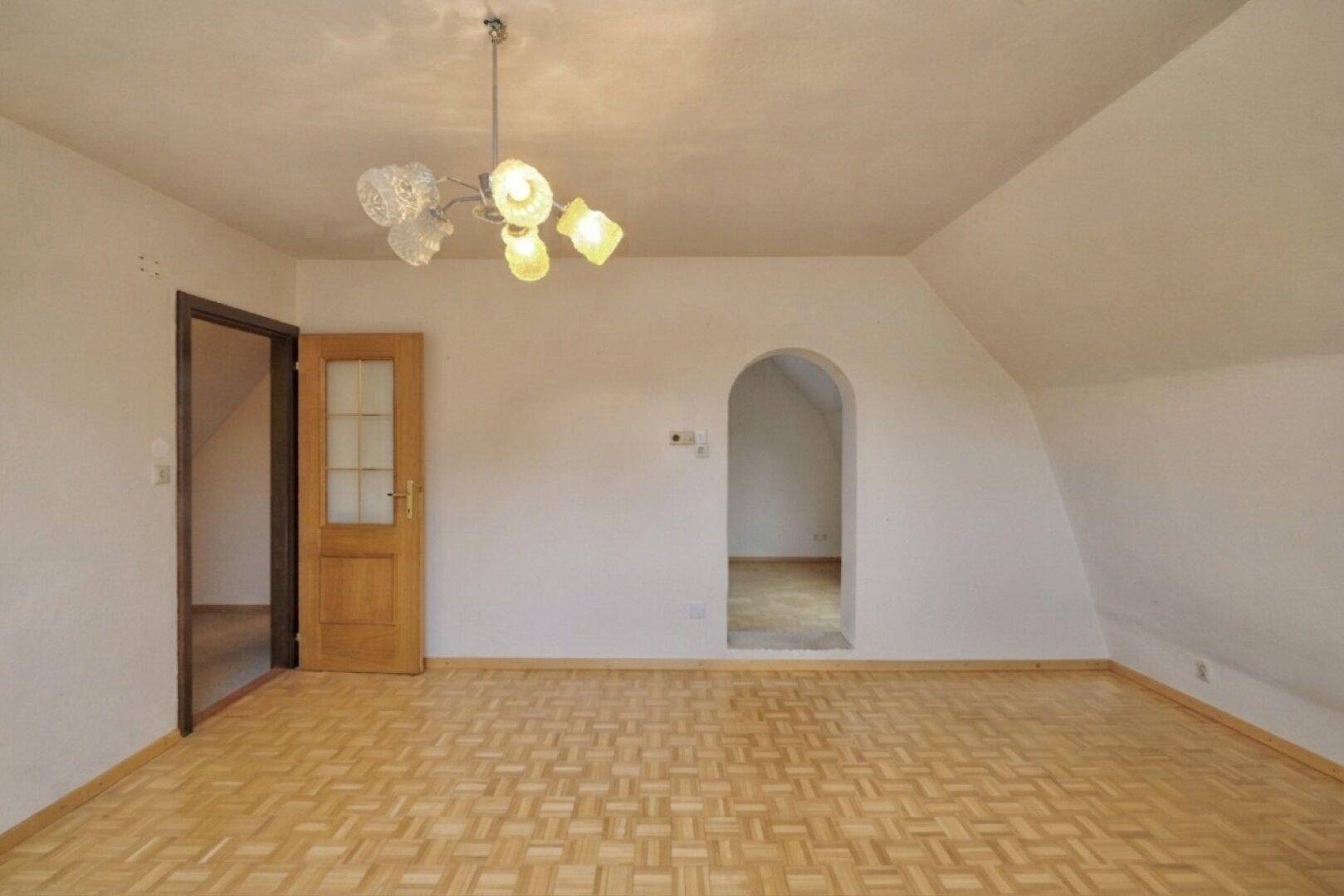 Heller Dachgeschossraum mit Parkettboden, Holztür und einem Bogen zu einem weiteren Bereich.