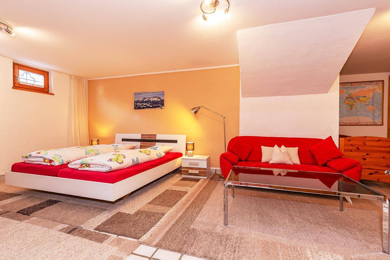 Schlafzimmer mit Doppelbett, rotem Sofa und Teppichboden in warmen Farbtönen.