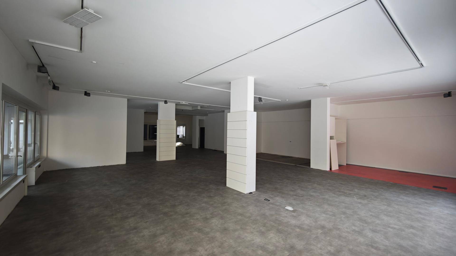 Weitläufiger, heller Geschäftsraum mit Betonboden, weißen Wänden und mehreren tragenden Säulen.