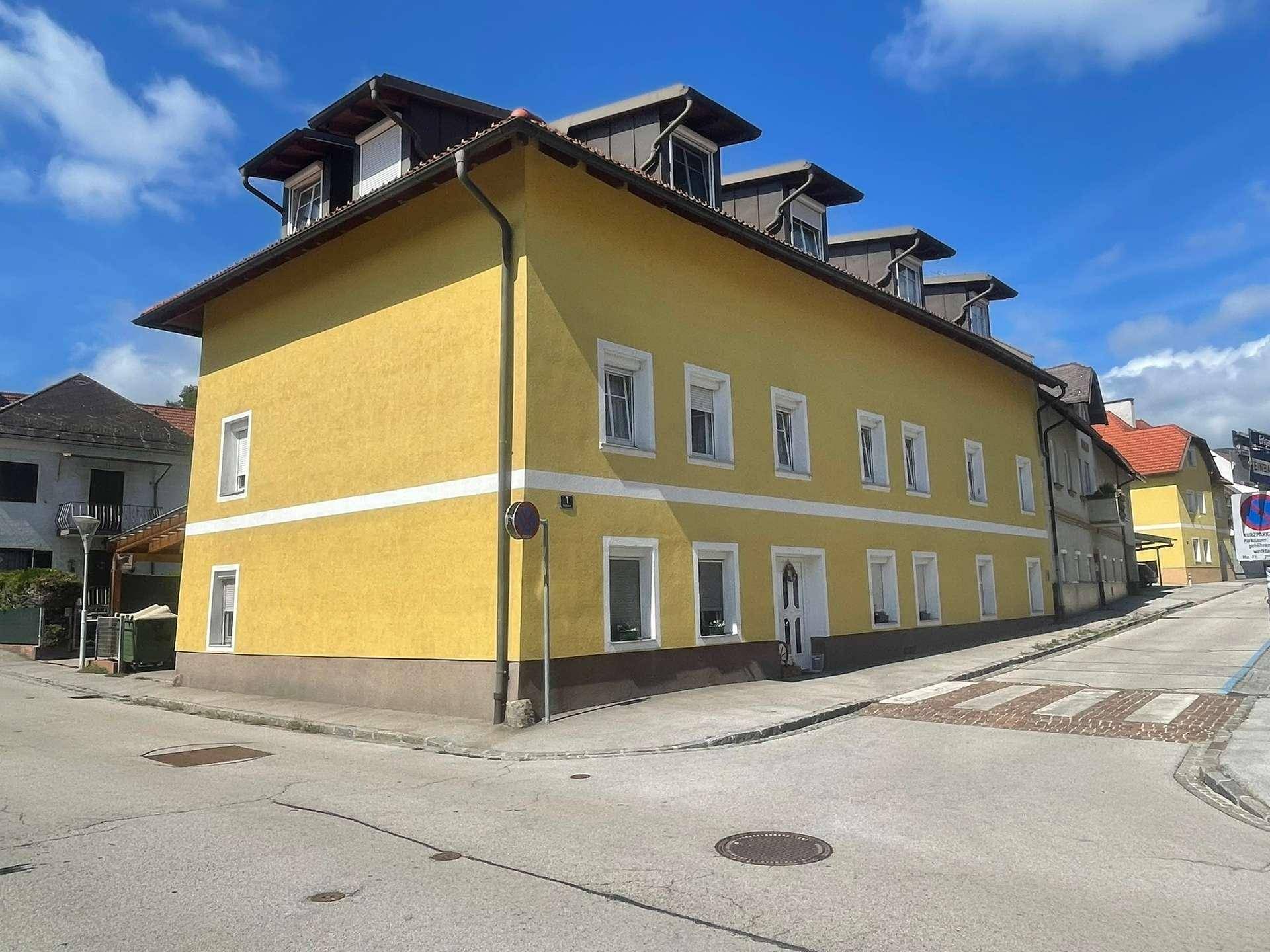 Eckgebäude mit gelber Fassade, weißen Fensterrahmen und Rollläden an einer Straße.