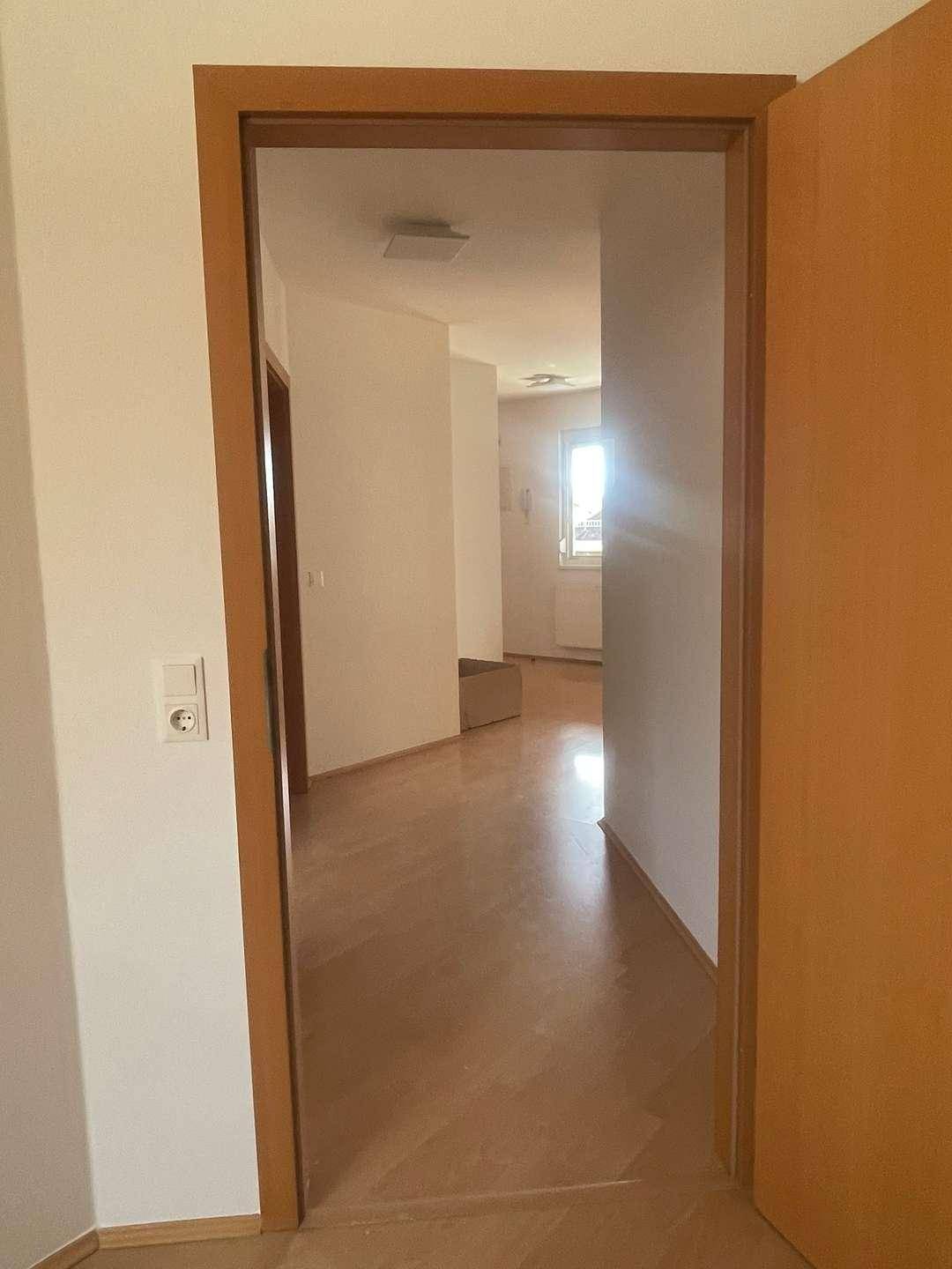 Blick durch eine offene Tür in einen hellen Flur mit Holzboden und Tageslicht.