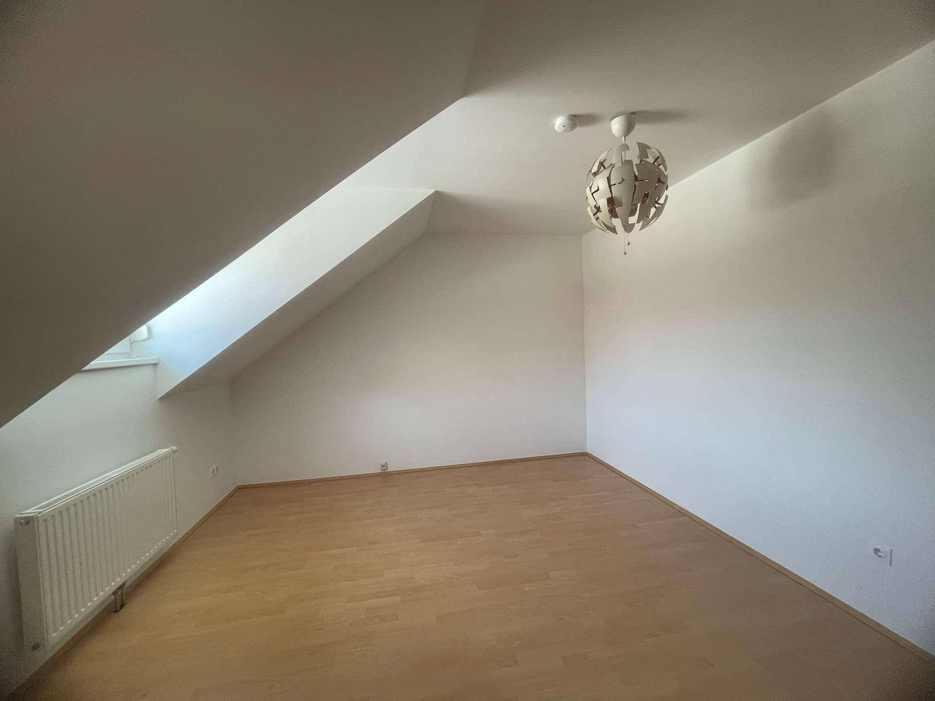 Helles Zimmer im Dachgeschoss mit schräger Decke, Dachfenster und Holzboden.