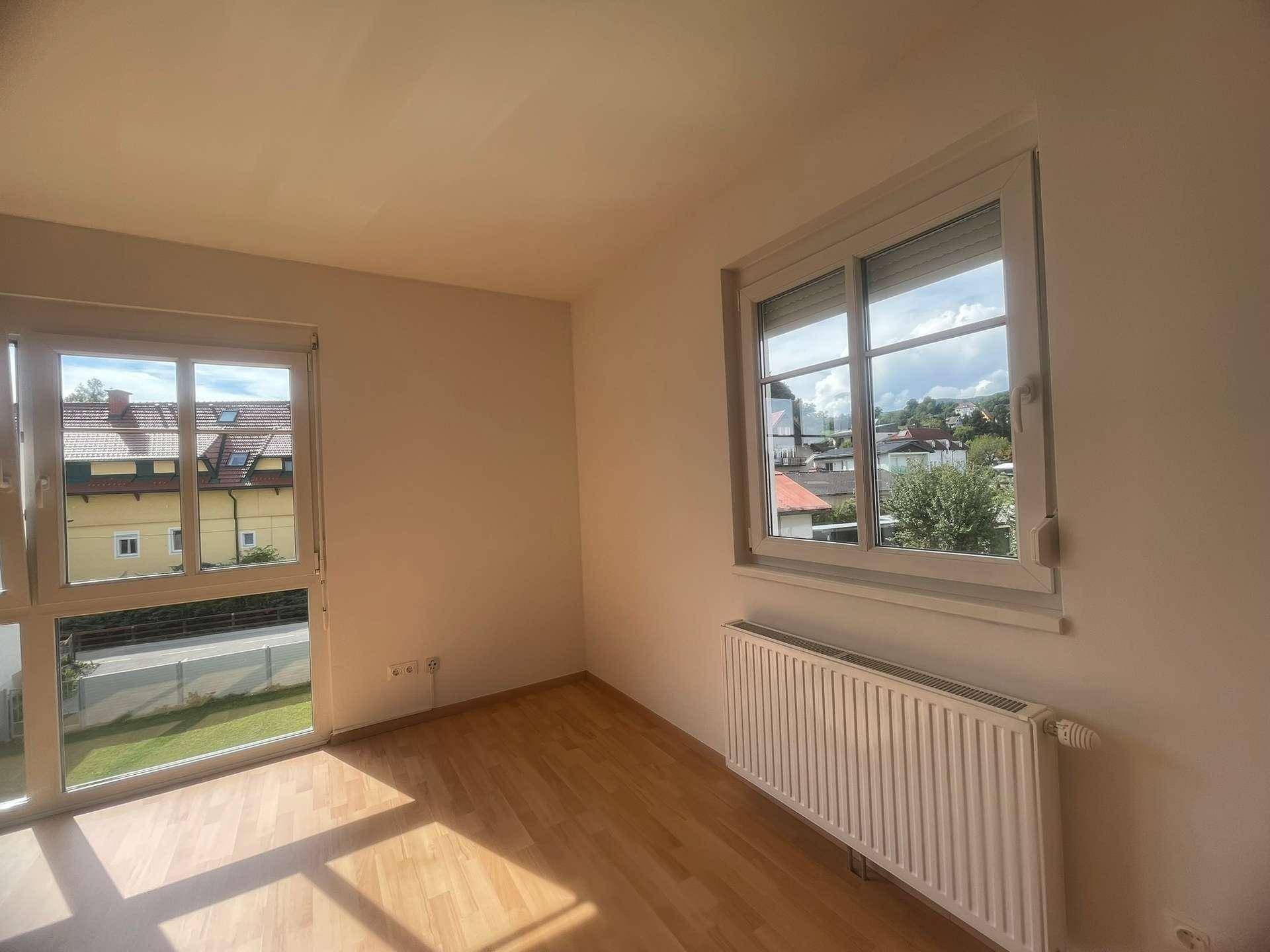 Helles Zimmer mit Holzboden und Fensterblick auf die grüne Umgebung und Häuser.