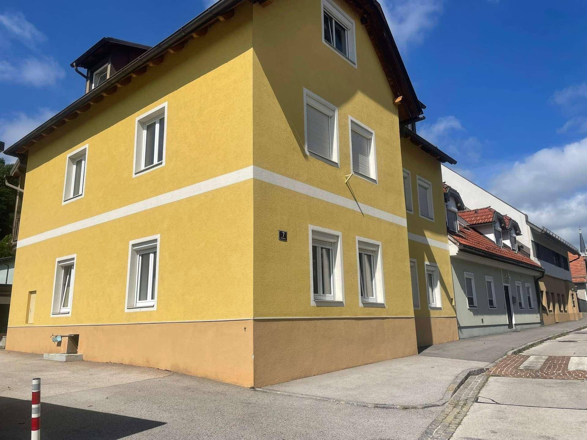Gelbe Hausfassade mit weißen Fensterrahmen und Rollläden an einer ruhigen Straße.
