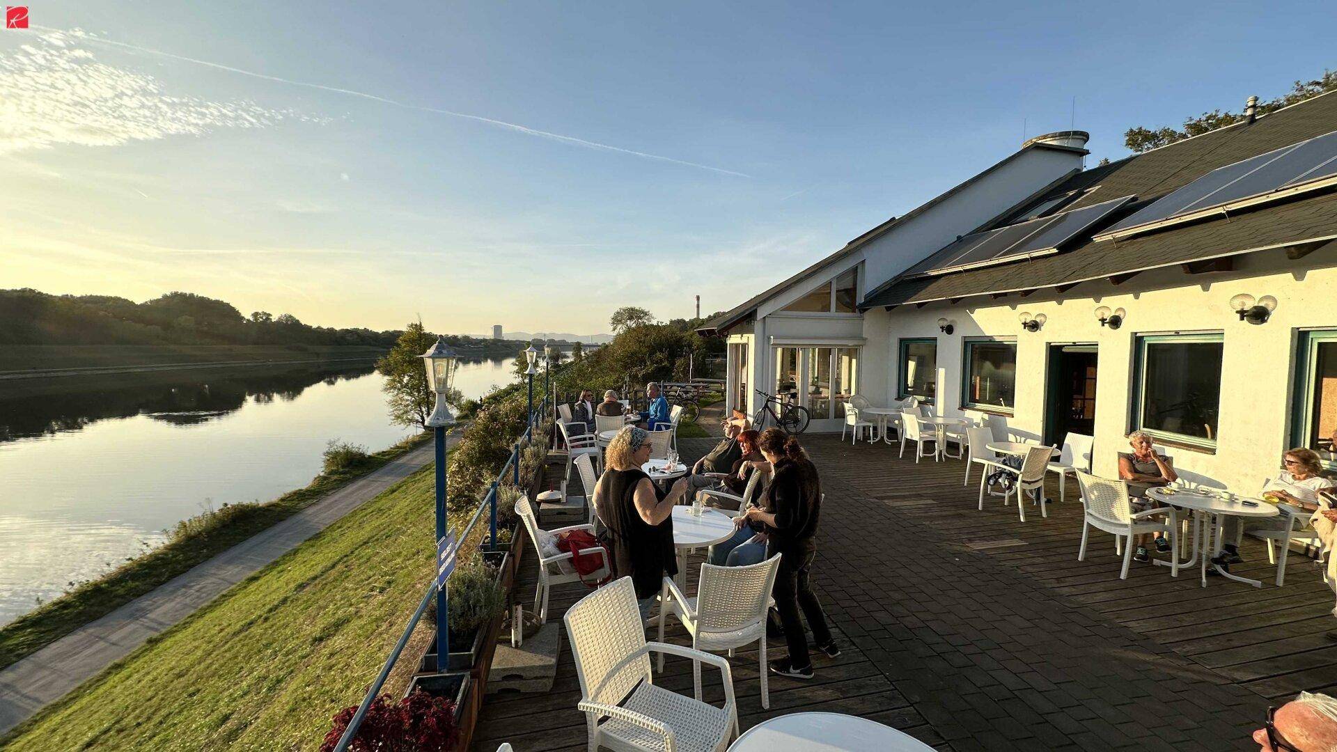Sonnige Außenterrasse eines Gebäudes mit Gästen, Blick auf den Fluss und Solaranlagen auf dem Dach.