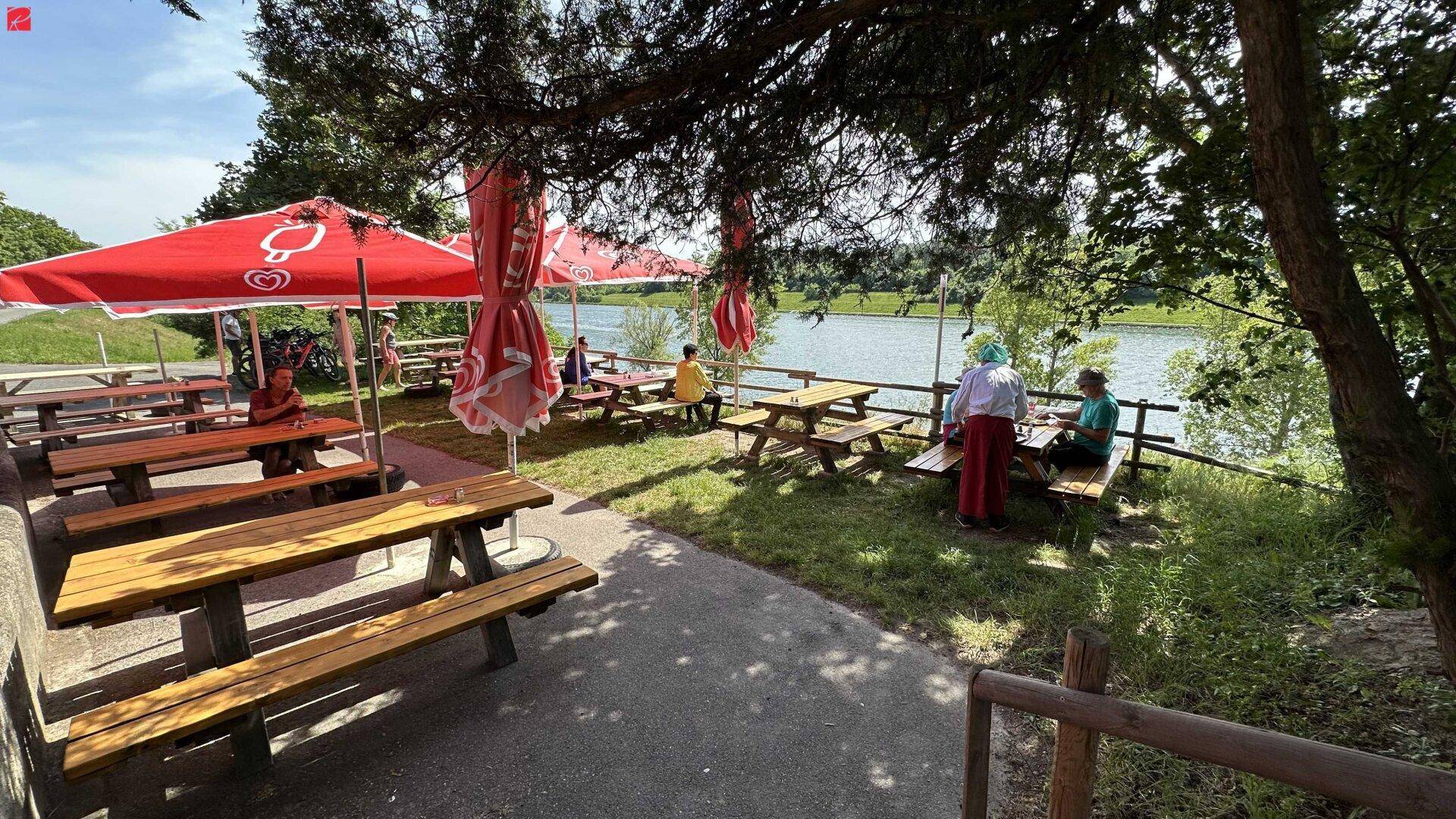Schattiger Picknickbereich im Freien mit Holztischen, roten Sonnenschirmen und Blick auf das Wasser.